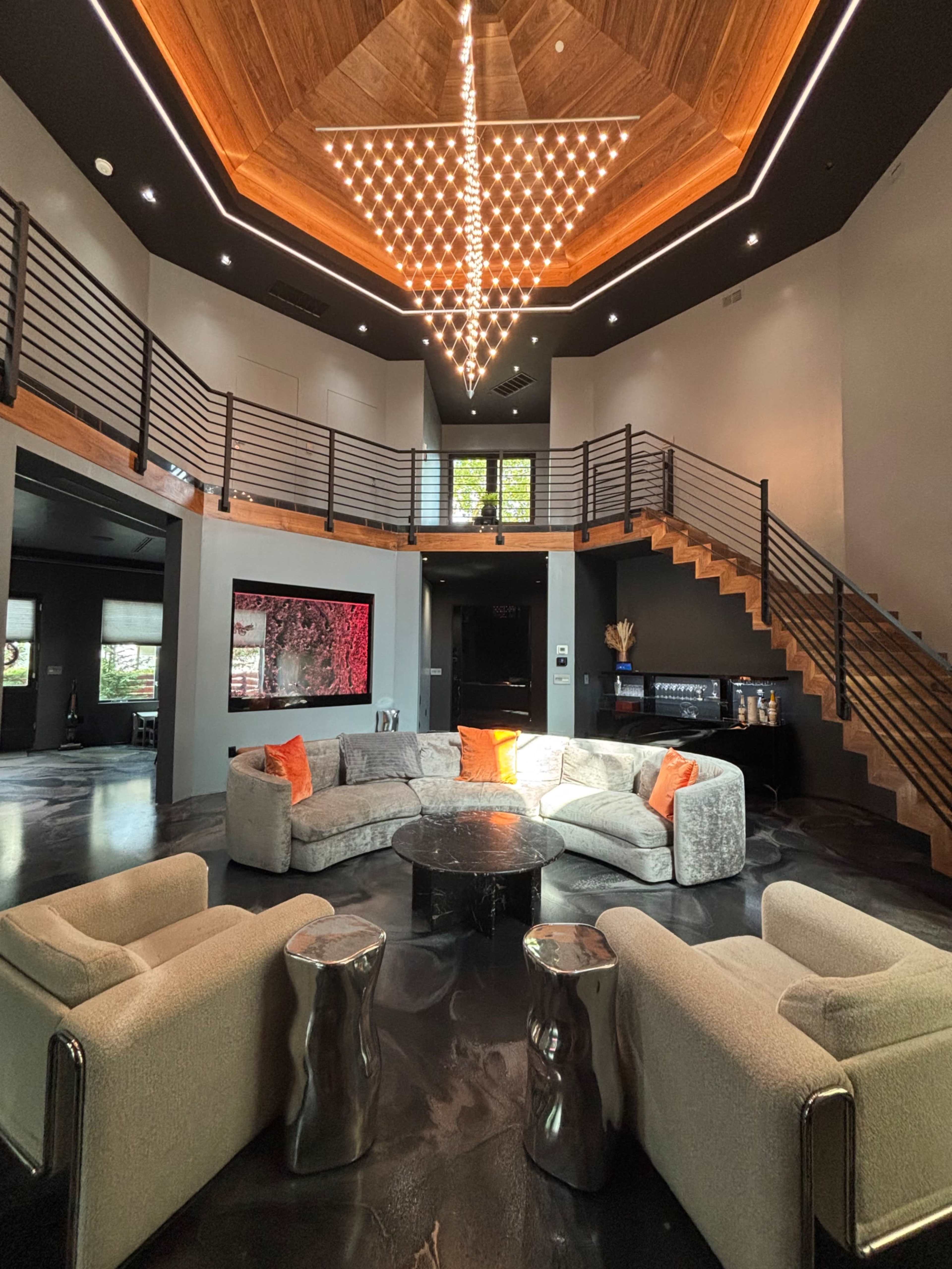 The image depicts a modern, upscale living room with a large circular sofa, stylish armchairs, and a dramatic chandelier above a sleek coffee table, surrounded by sleek wooden and glass features.