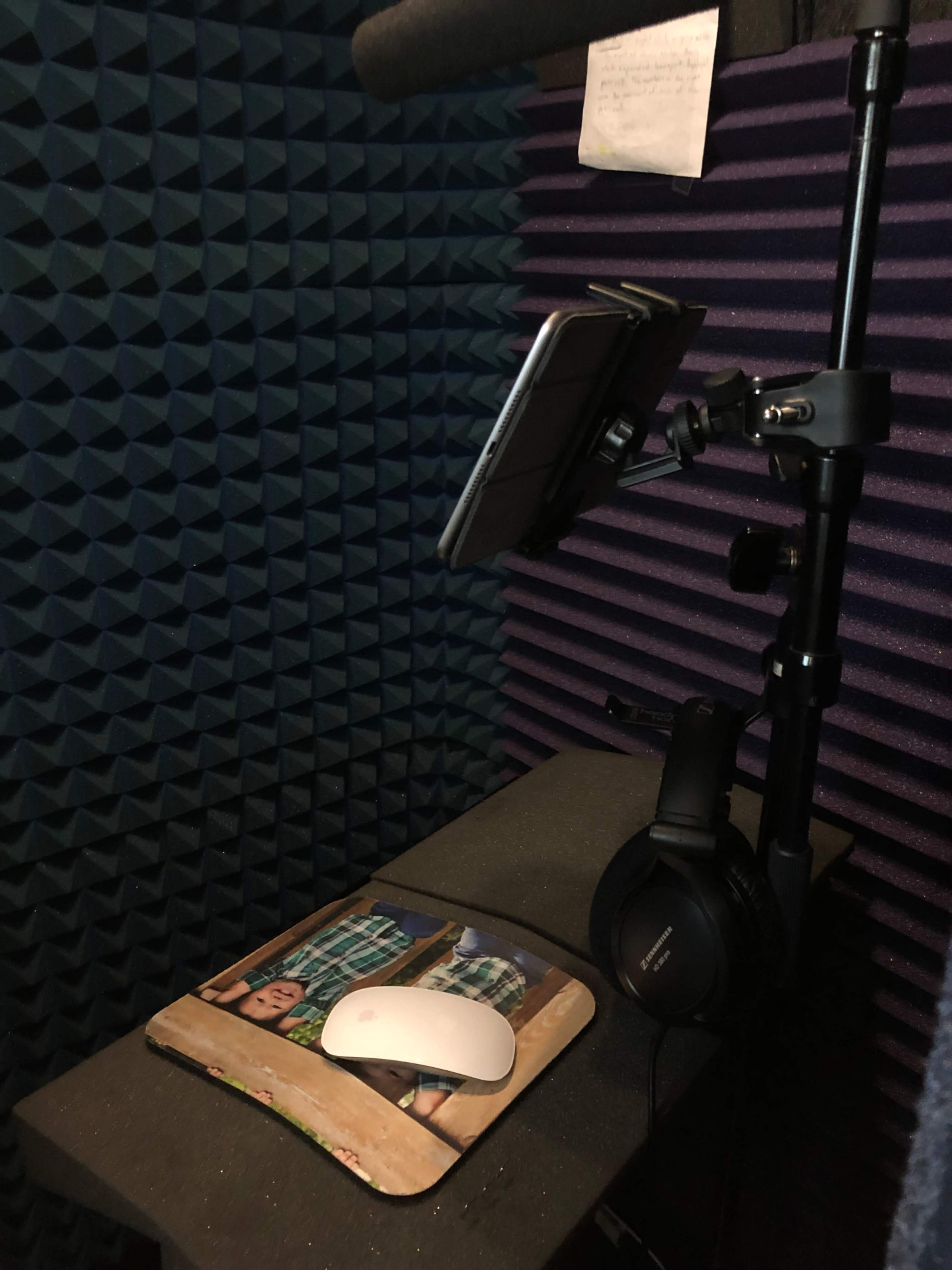 The image shows a recording booth lined with acoustic foam, featuring a microphone on a boom arm, a tablet holder, and a desk with a mouse and headphones.