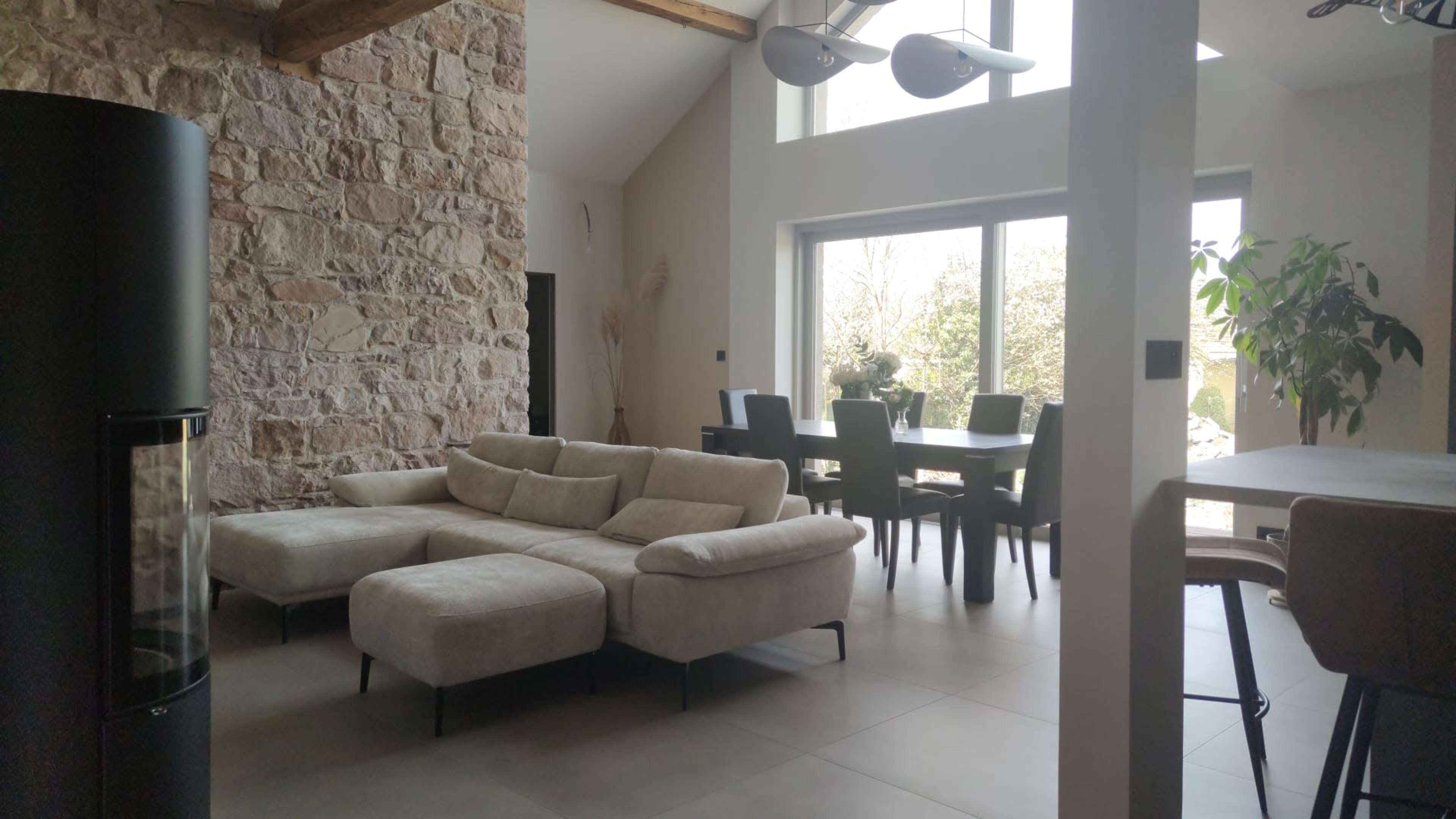 The image shows a modern living room with a light-colored sectional sofa, stone walls, and a dining area featuring a large table and dark chairs under bright natural light from large windows.