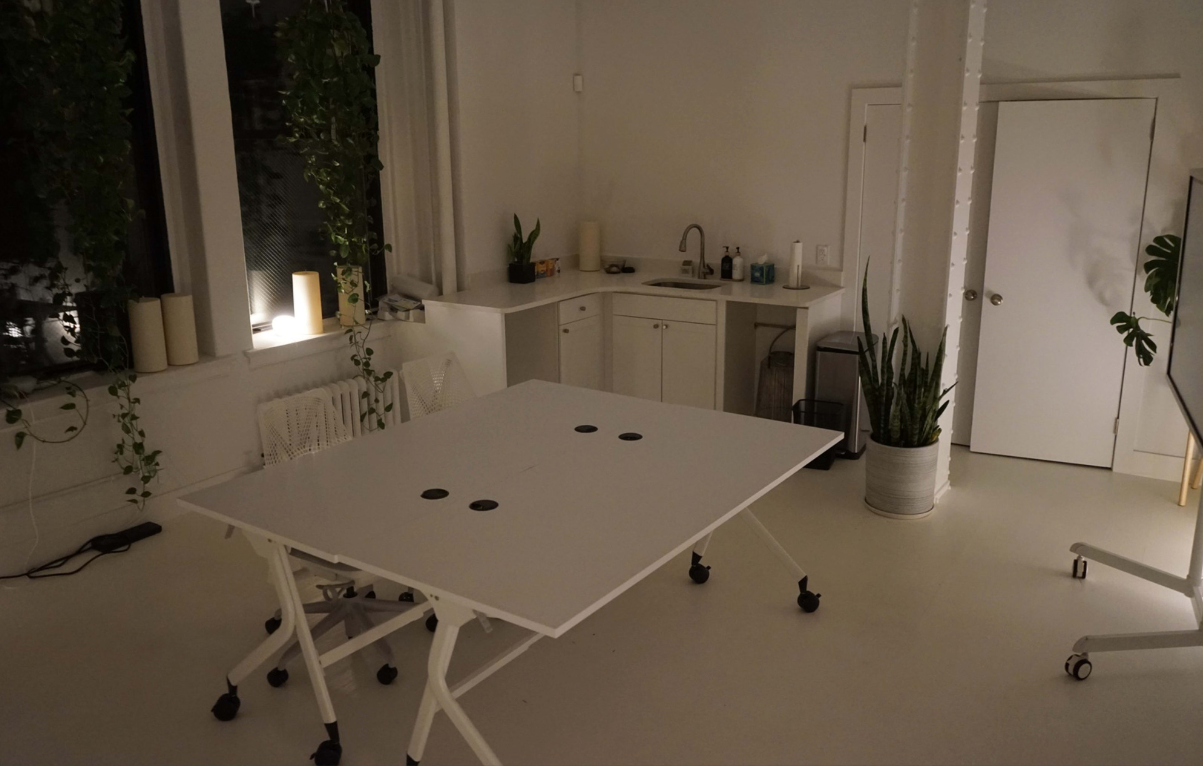 The image shows a minimalist office space featuring a white folding table, a kitchenette, and several indoor plants.
