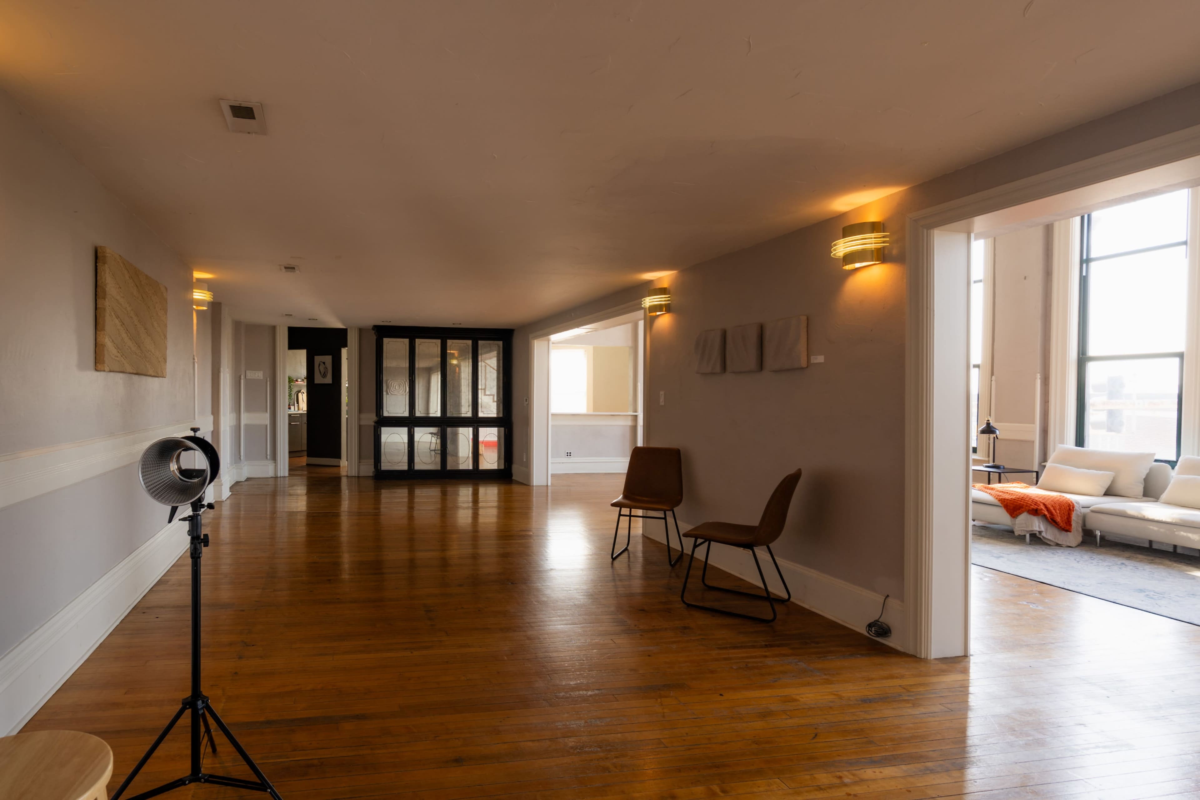 The image shows a spacious interior hallway with wooden floors, featuring a standing fan, a chair, and large windows allowing natural light to enter.