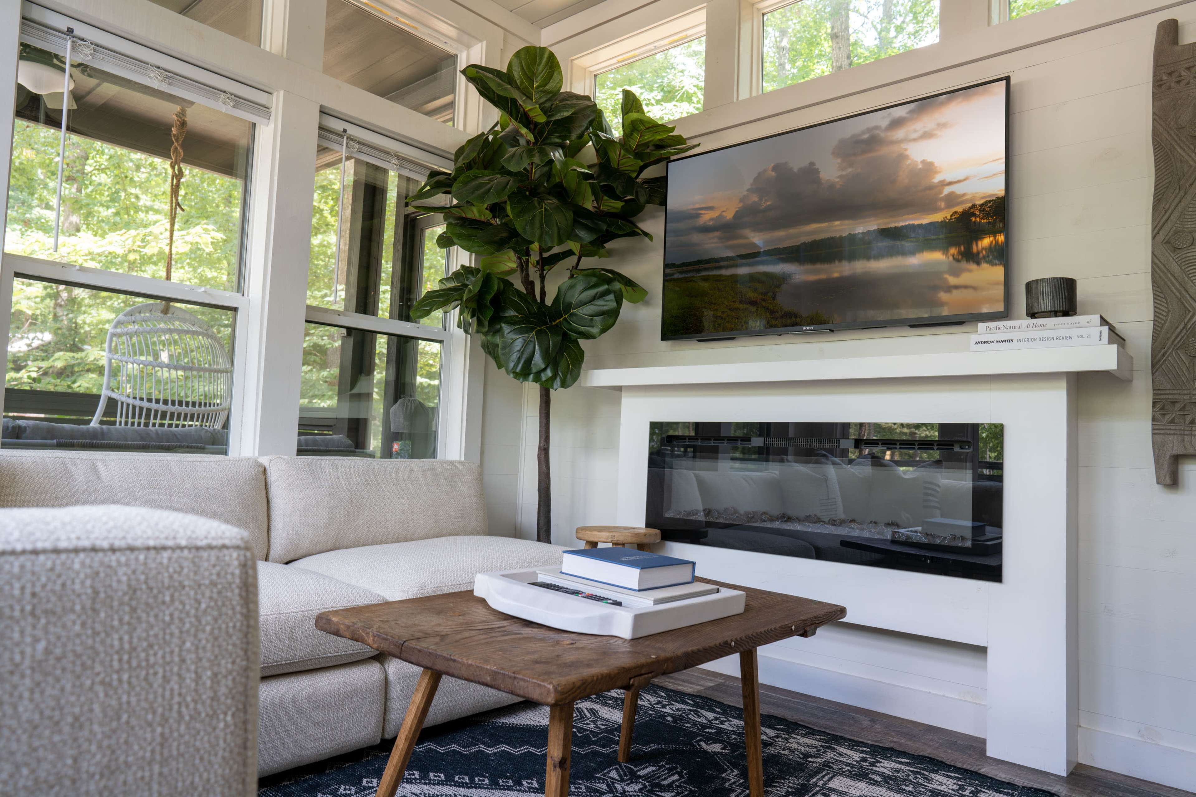 A cozy living room with a beige sectional sofa, a wooden coffee table, an electric fireplace, and a large wall-mounted television above a white mantle, all set against windows that overlook greenery.