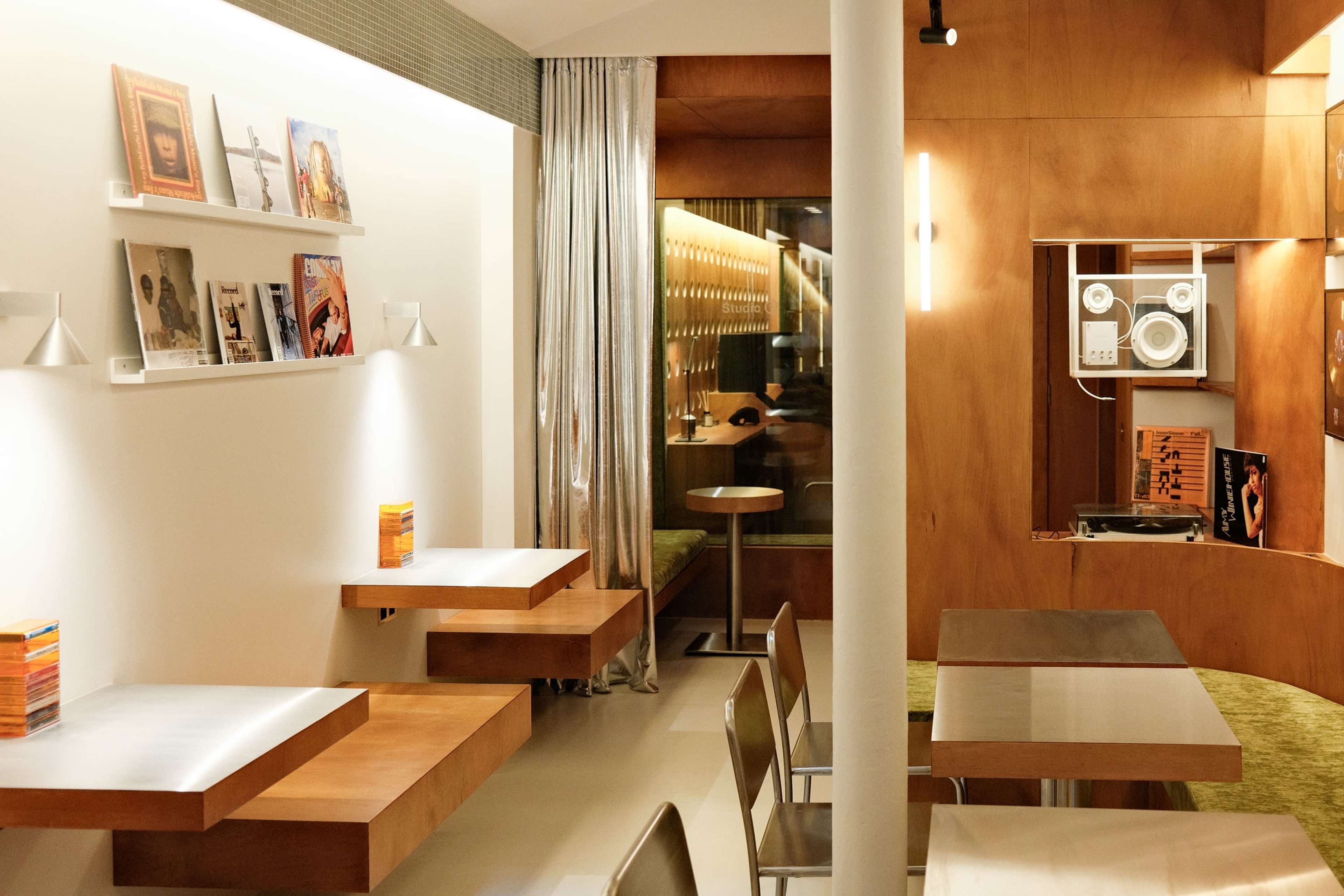 The image shows a modern cafe with wooden seating, a record player, and shelves displaying books and magazines.