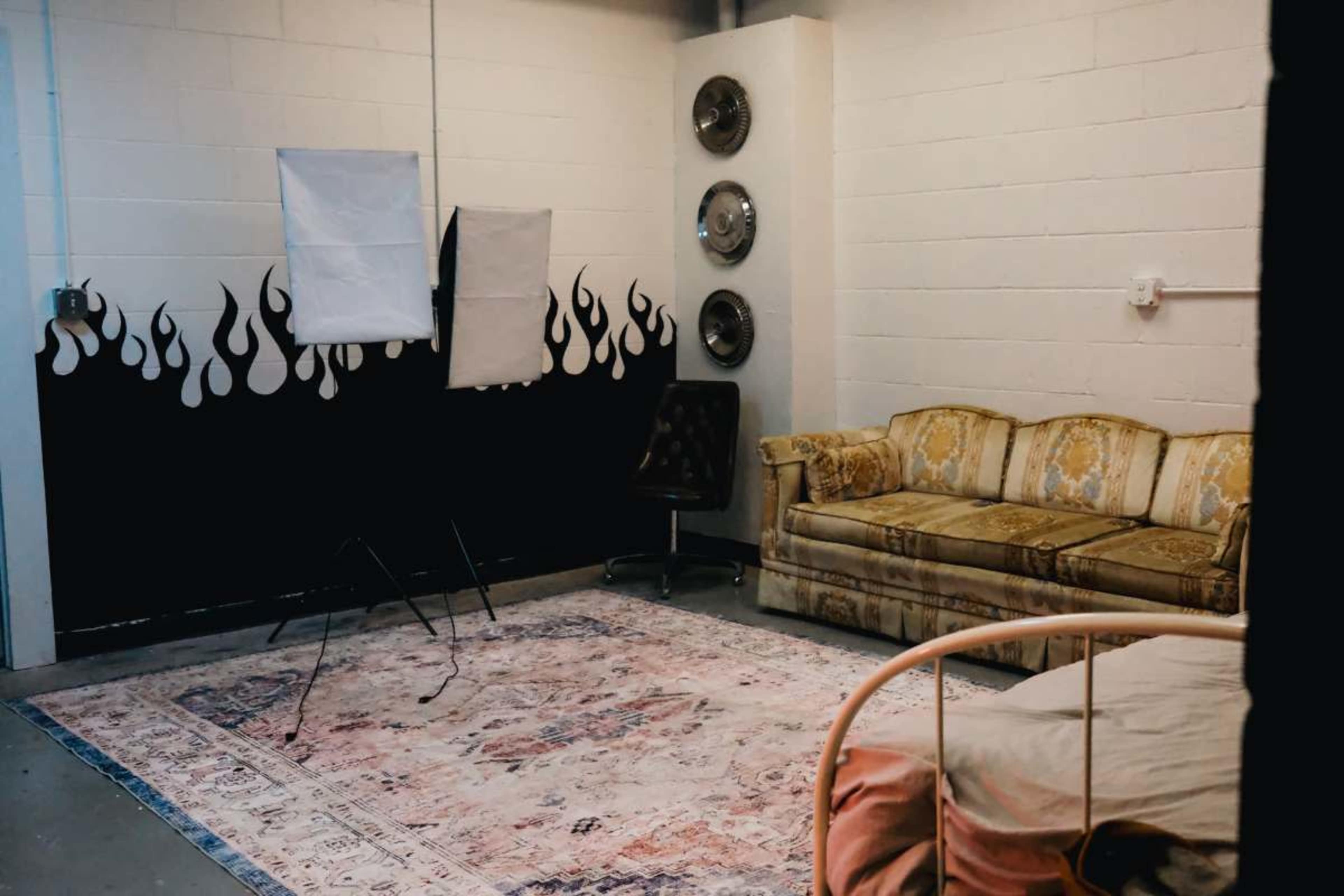The scene features a corner of a room with a patterned rug, an ornate couch, a bed, and two lighting setup stands against a wall decorated with flame motifs.