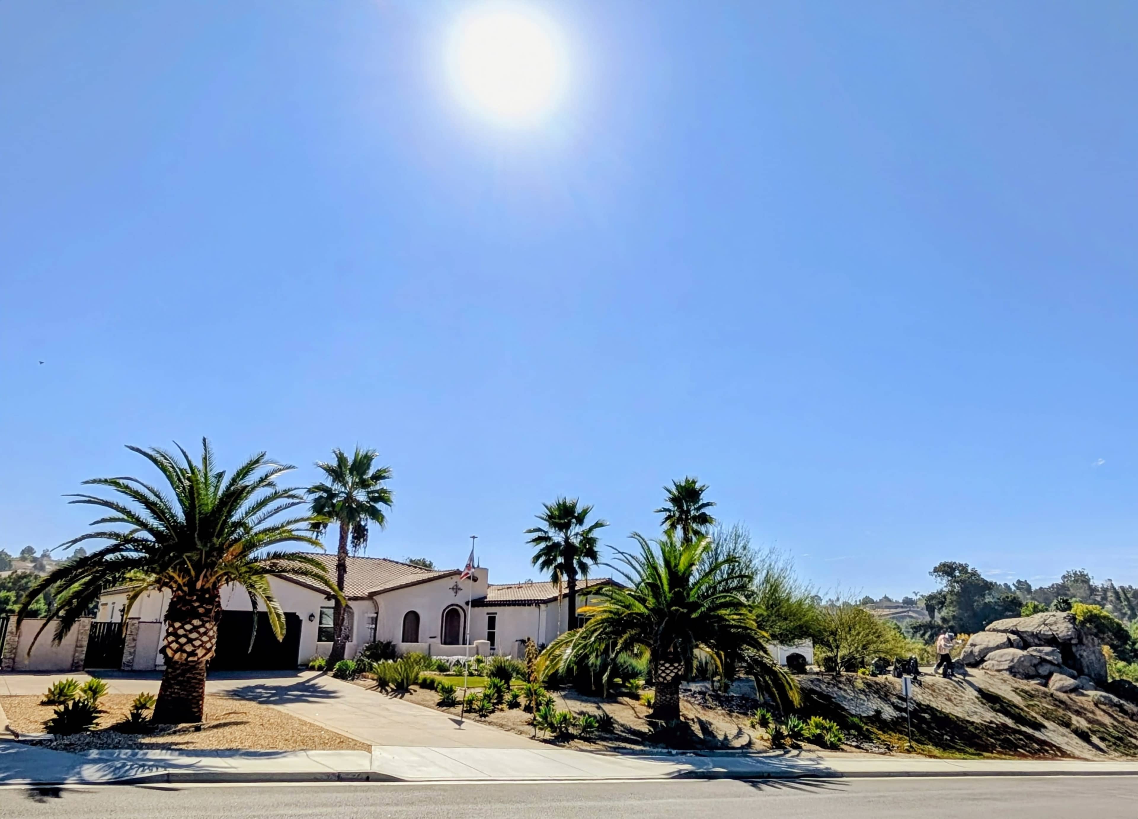 A single-story house with a stucco exterior and a red tile roof is surrounded by palm trees under a clear blue sky.