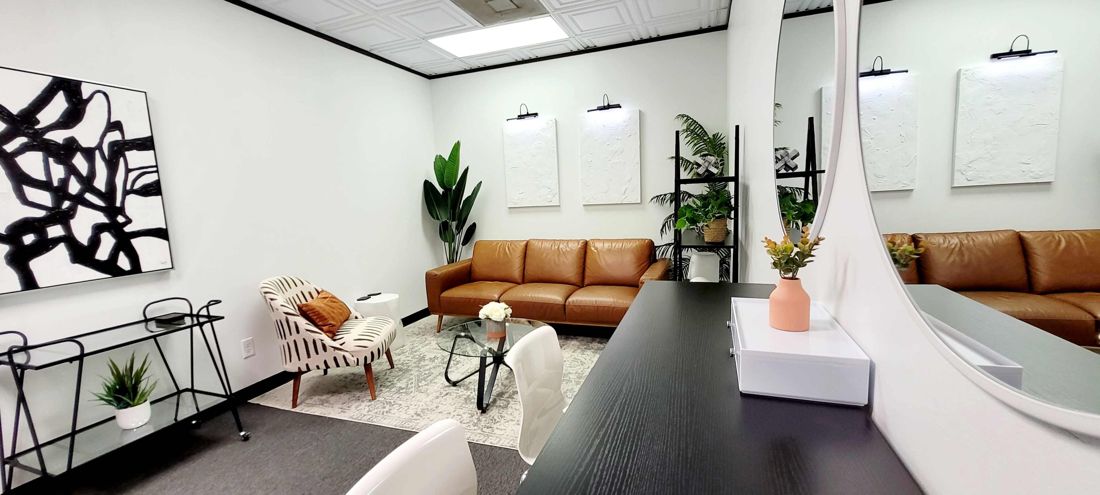 A modern office space featuring a brown leather sofa, a patterned chair, a glass coffee table, and potted plants, with white walls adorned by artwork and a mirror.
