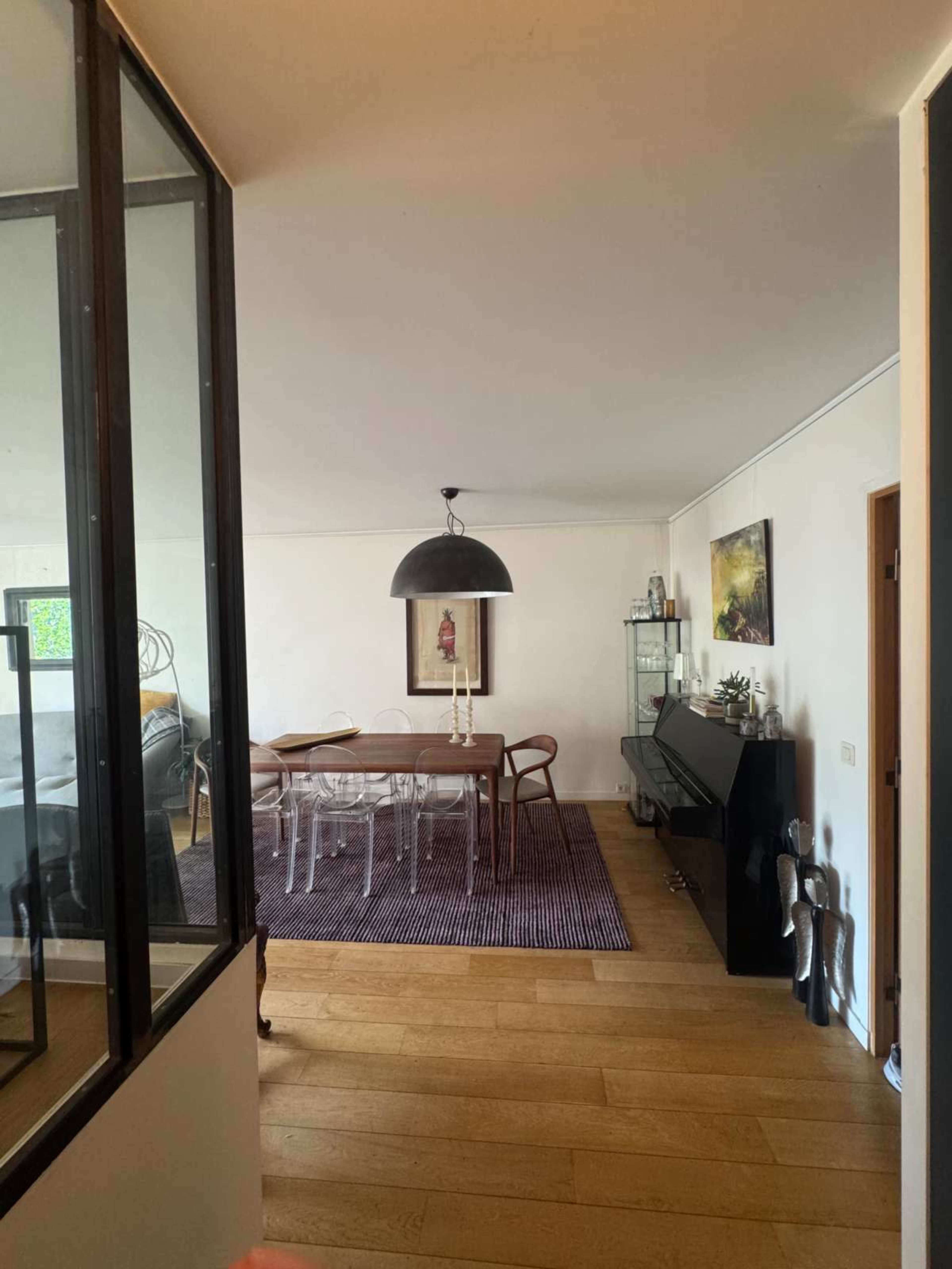The image shows a modern interior space featuring a dining area with a wooden table, transparent chairs, a large pendant light, and a dark piano, all set against light-colored walls and wooden flooring.