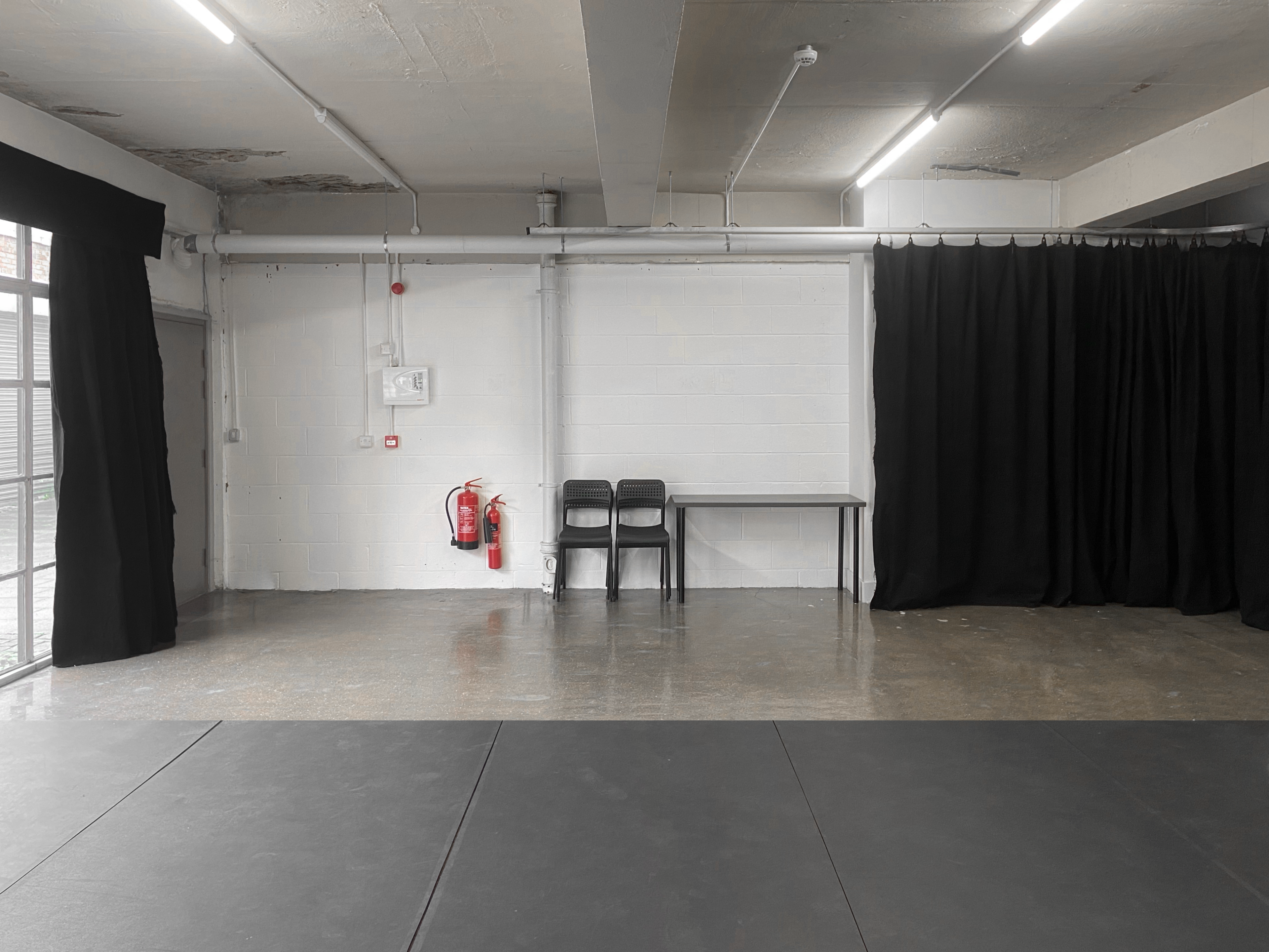The scene shows a minimalist room with a concrete floor, two black chairs, a table, a fire extinguisher mounted on the wall, and black curtains along one side.