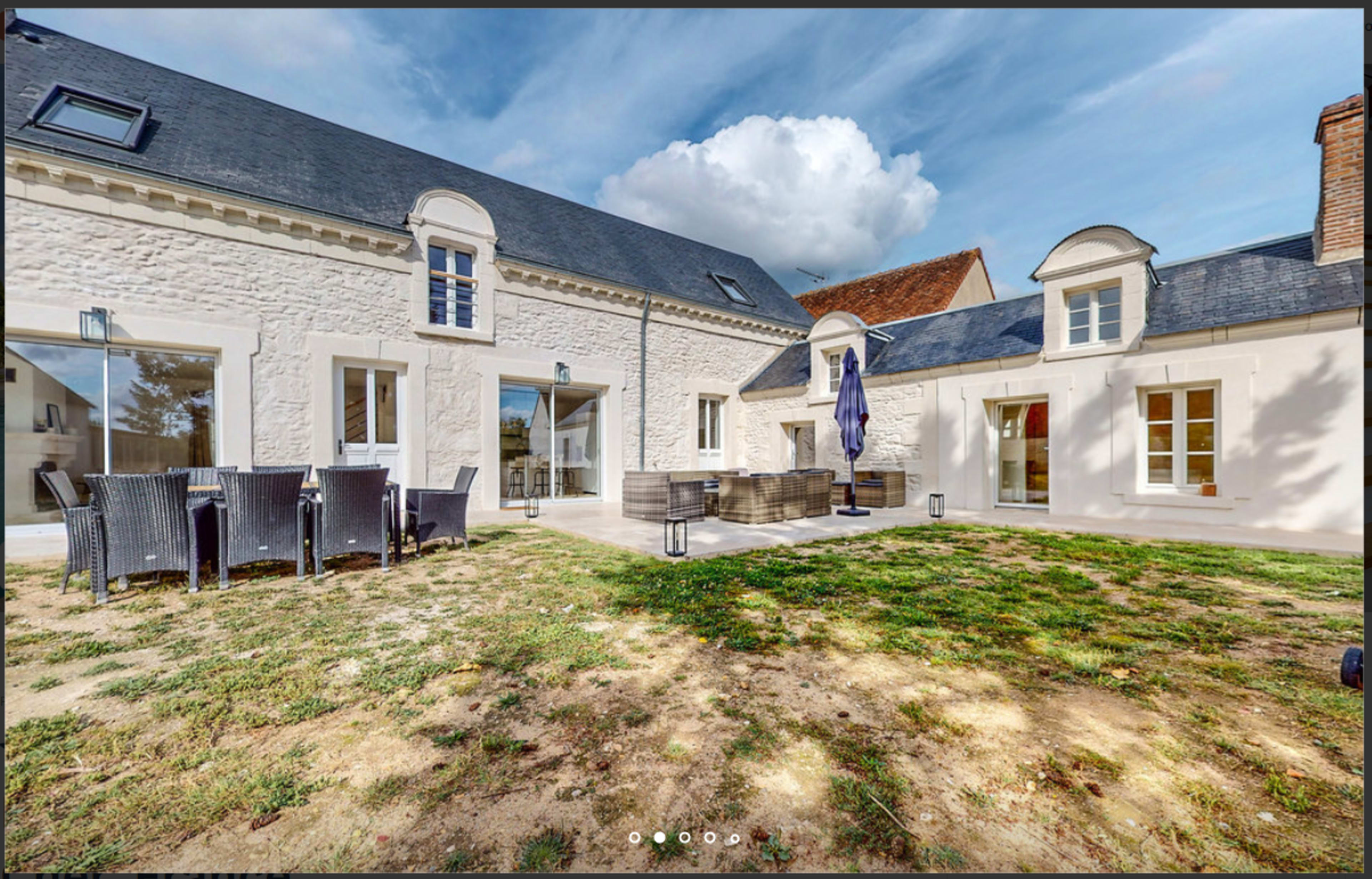 The image shows a modern courtyard with a stone-clad house, outdoor seating area, and a grassy yard under a partly cloudy sky.