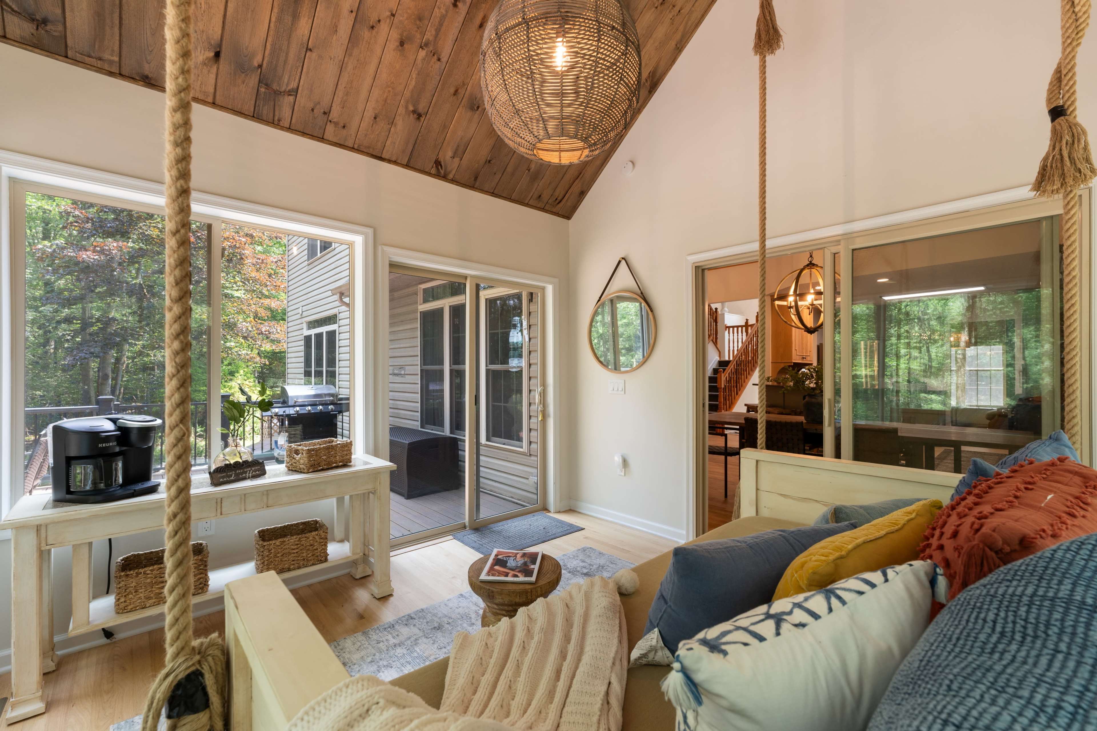 The image shows a bright sunroom with a wooden ceiling, featuring a cozy seating area with colorful pillows and a view of a green outdoor space through large windows.