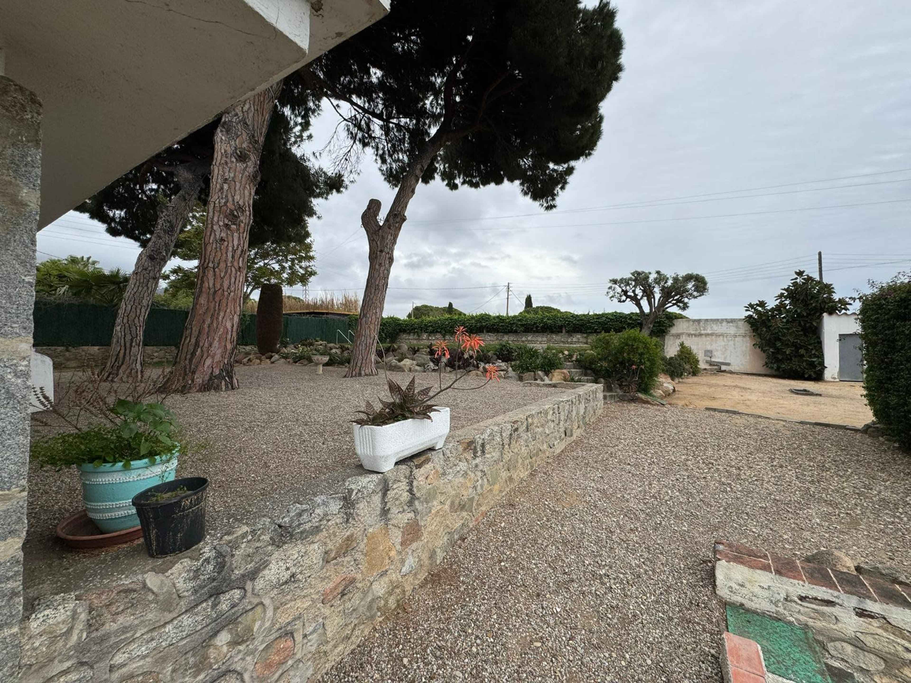 The image shows a gravel garden area surrounded by stone walls, featuring potted plants and trees, with a cloudy sky overhead.