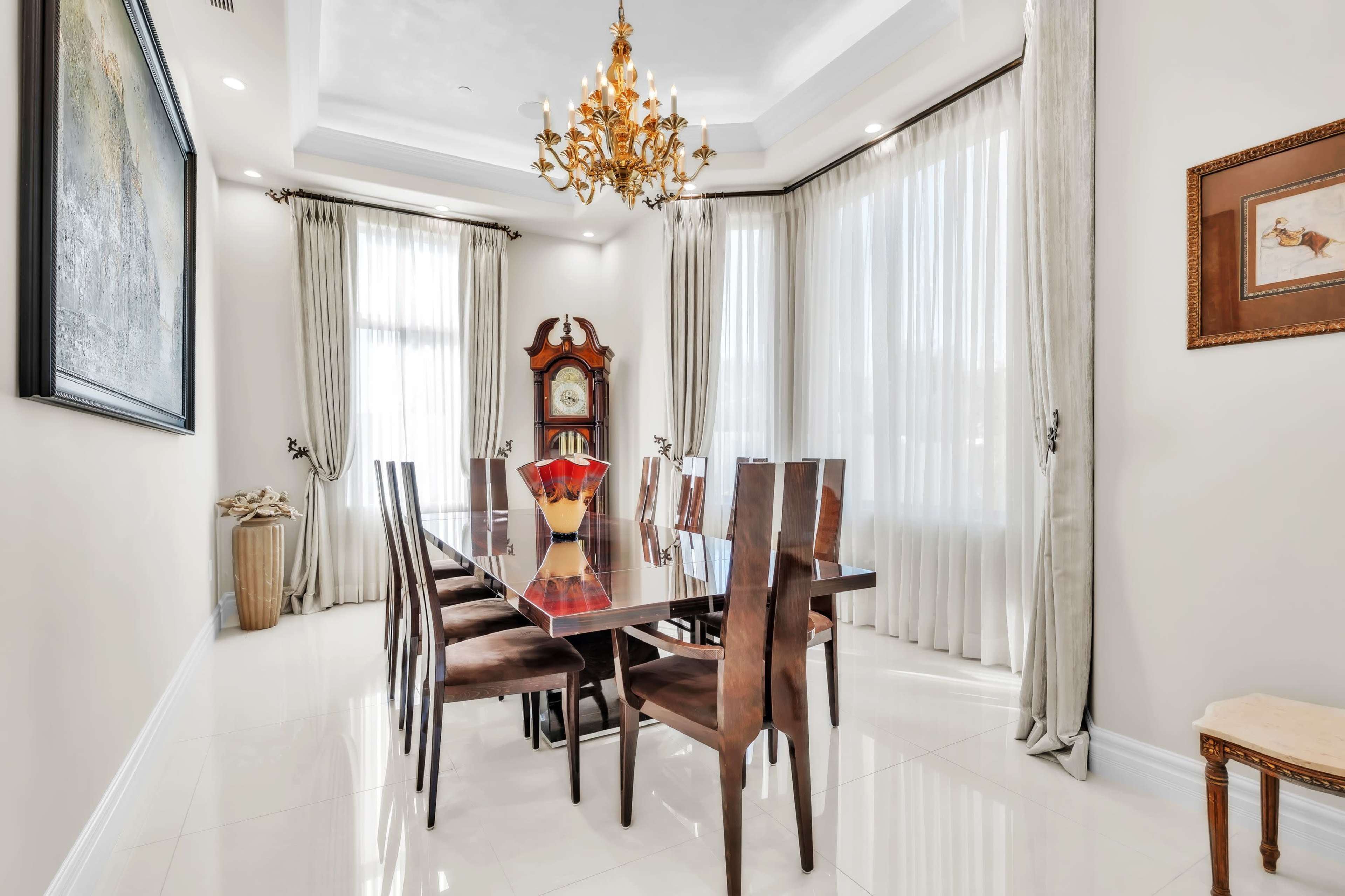A dining room features a long glass table surrounded by eight chairs, with a vintage clock and decorative items placed around large windows partially covered by drapes.