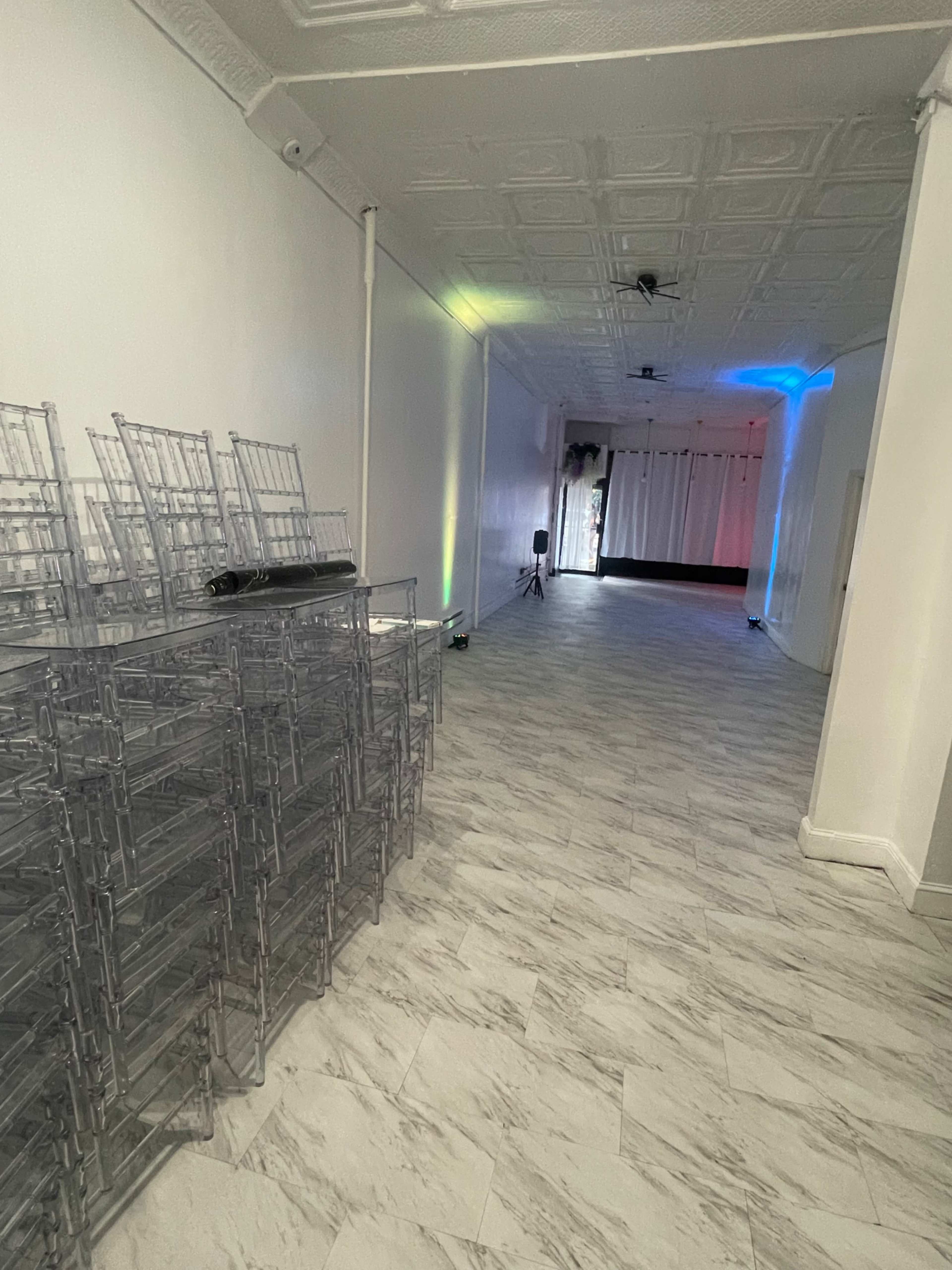 The image shows a long, narrow hallway with a stack of clear chairs on the left and a brightly lit event space in the distance.