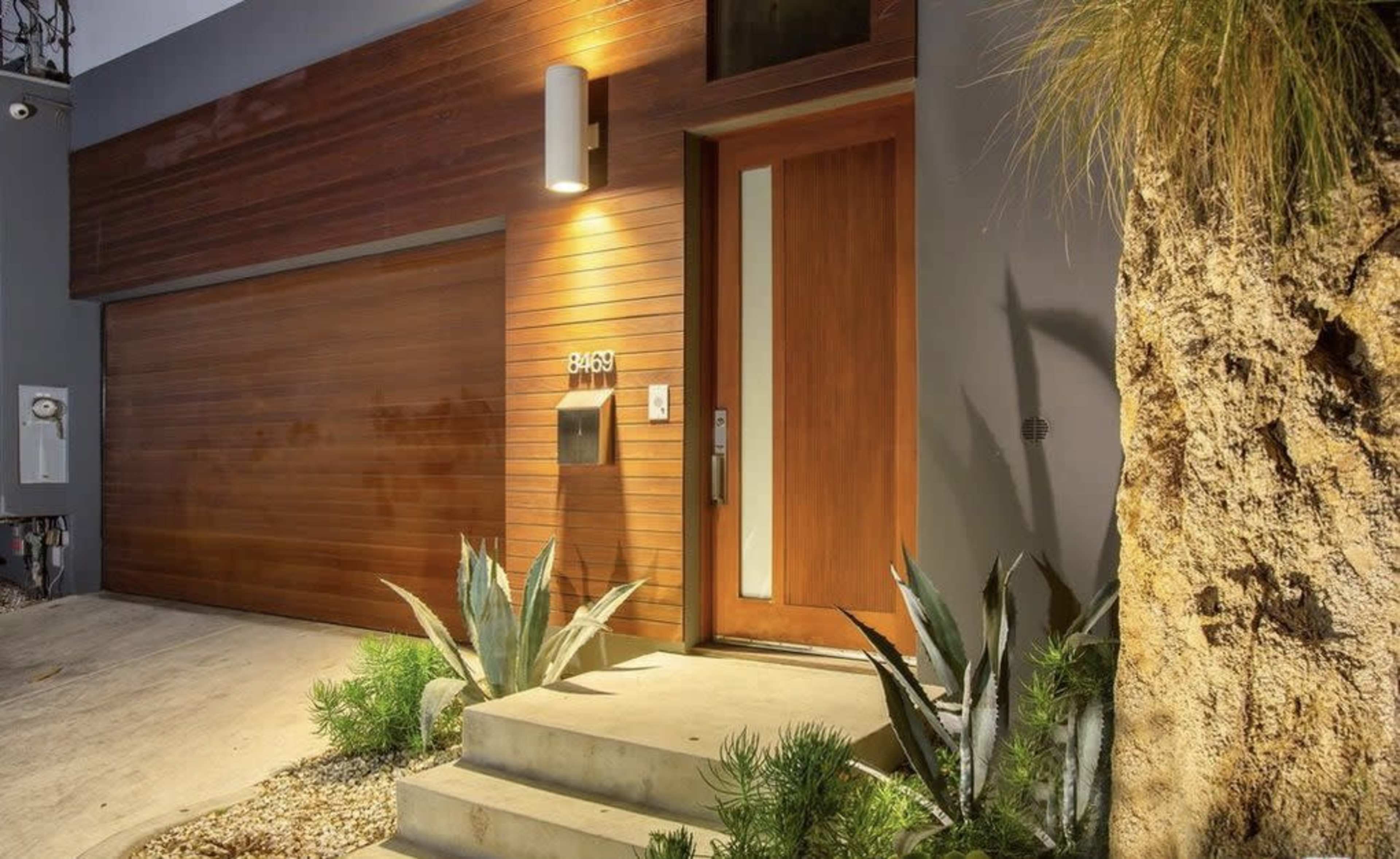 The image shows a modern entrance of a house with a wooden door and a garage door, accompanied by plants and a stone pathway.