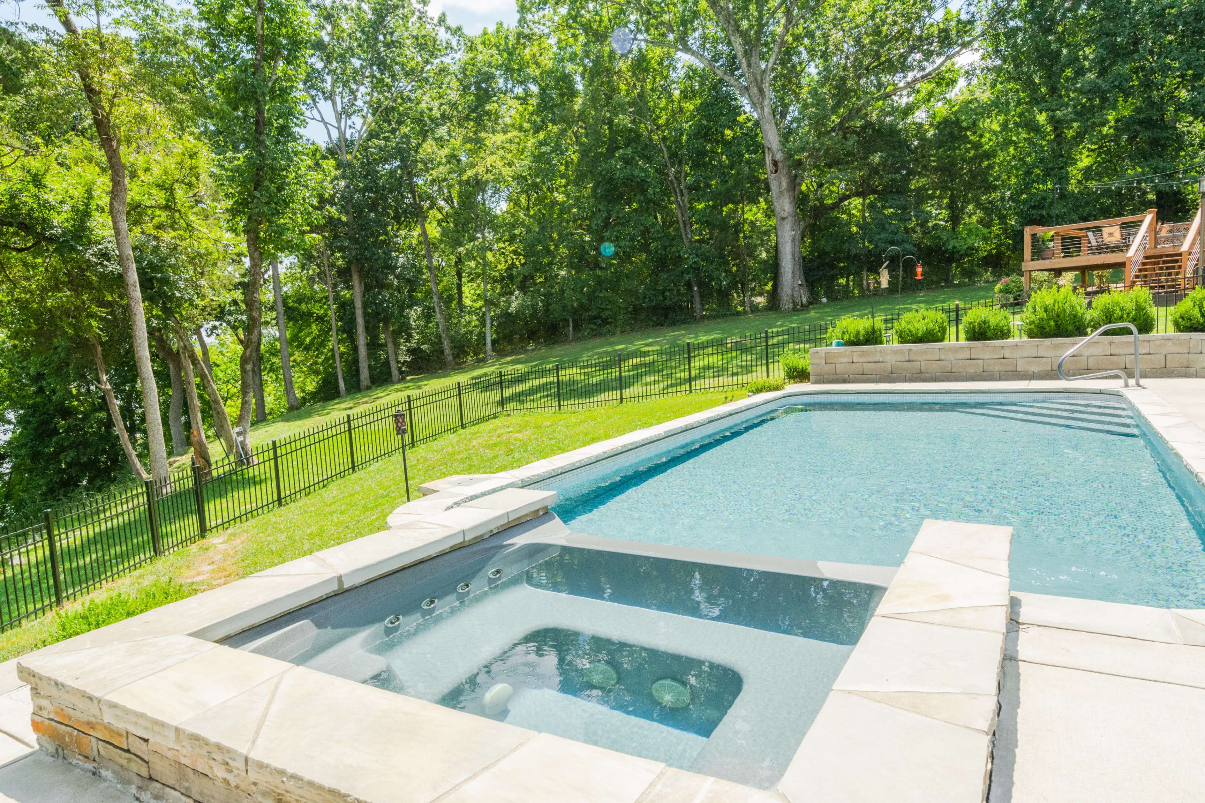 A backyard with a swimming pool and a spa area, surrounded by a fenced lawn and trees.