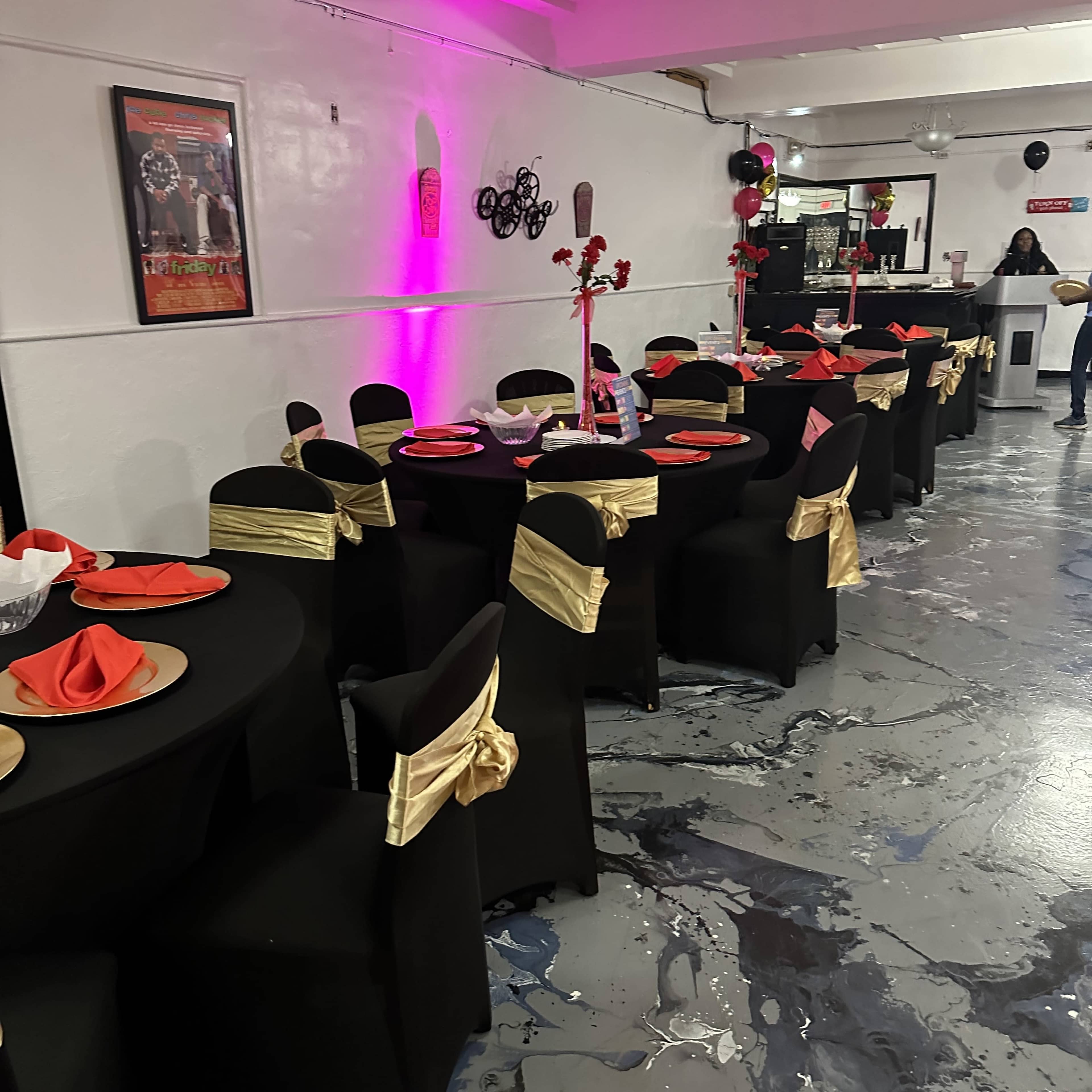 A banquet hall is set up with black tablecloths, gold sashes on chairs, and red napkins on the tables, illuminated by purple lighting.