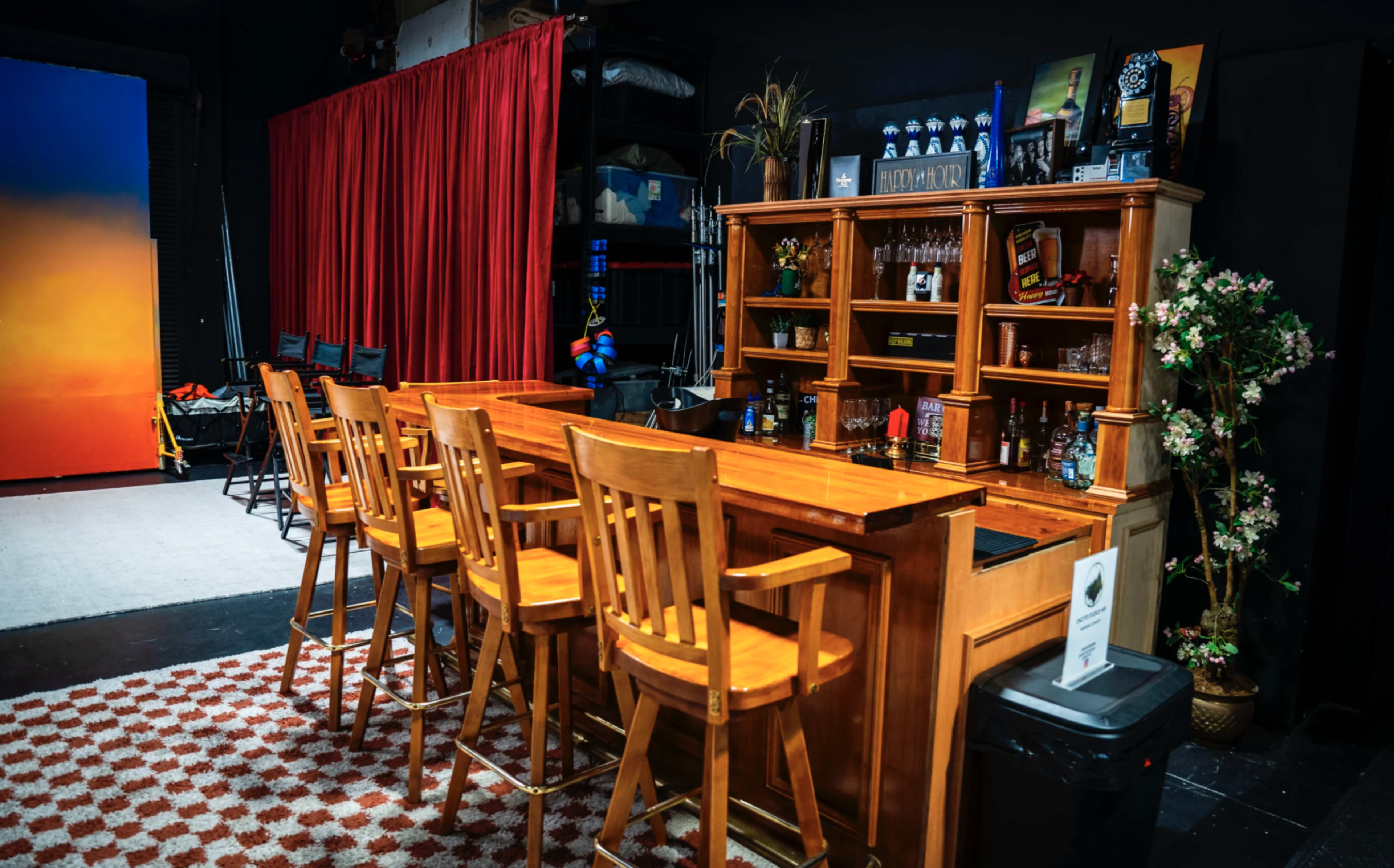 The image shows a wooden bar with high stools, set against a backdrop of red curtains and shelves stocked with various liquor bottles.