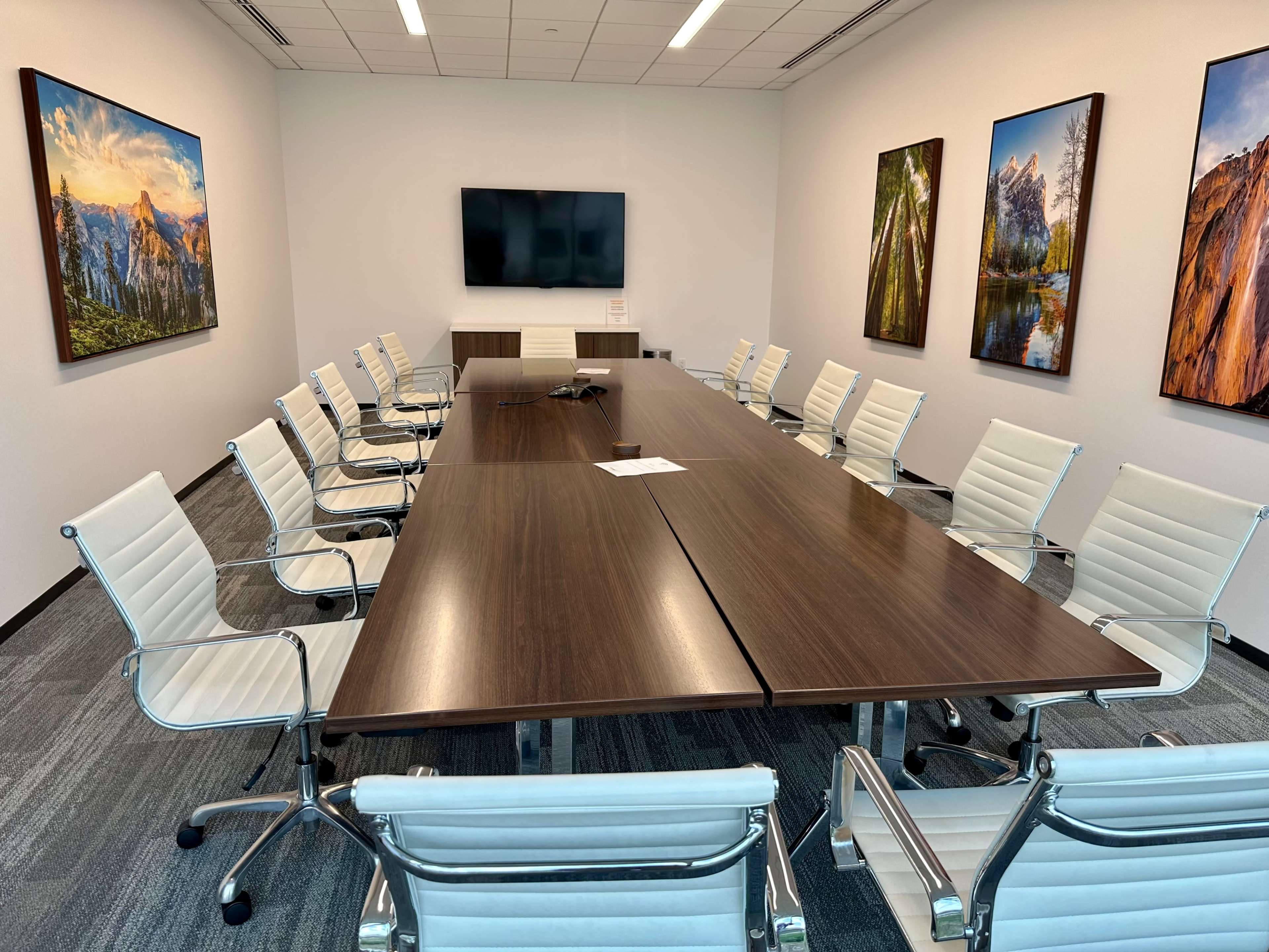 A modern conference room with a large wooden table surrounded by white chairs, featuring wall-mounted artwork and a television screen.