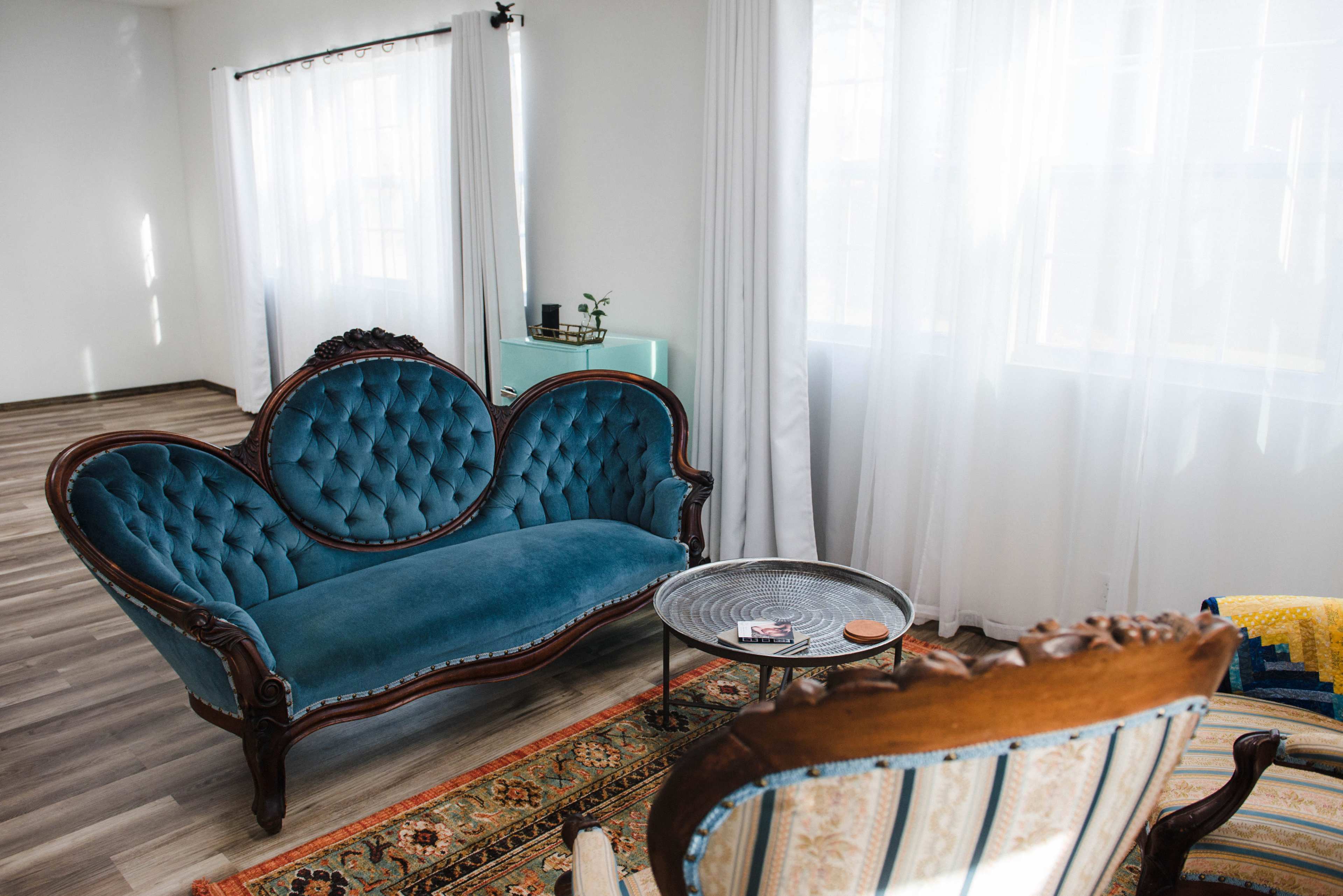 A vintage-style living room features a teal velvet sofa and a patterned area rug, with natural light filtering through sheer white curtains.