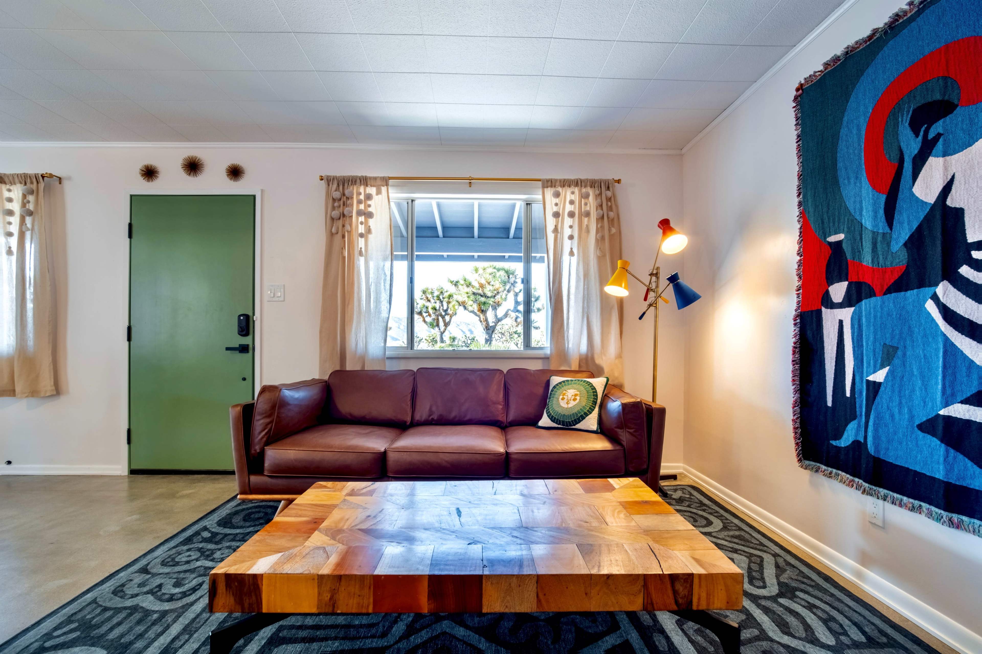A living room with a brown leather couch, a wooden coffee table, and a colorful wall hanging in a well-lit space.