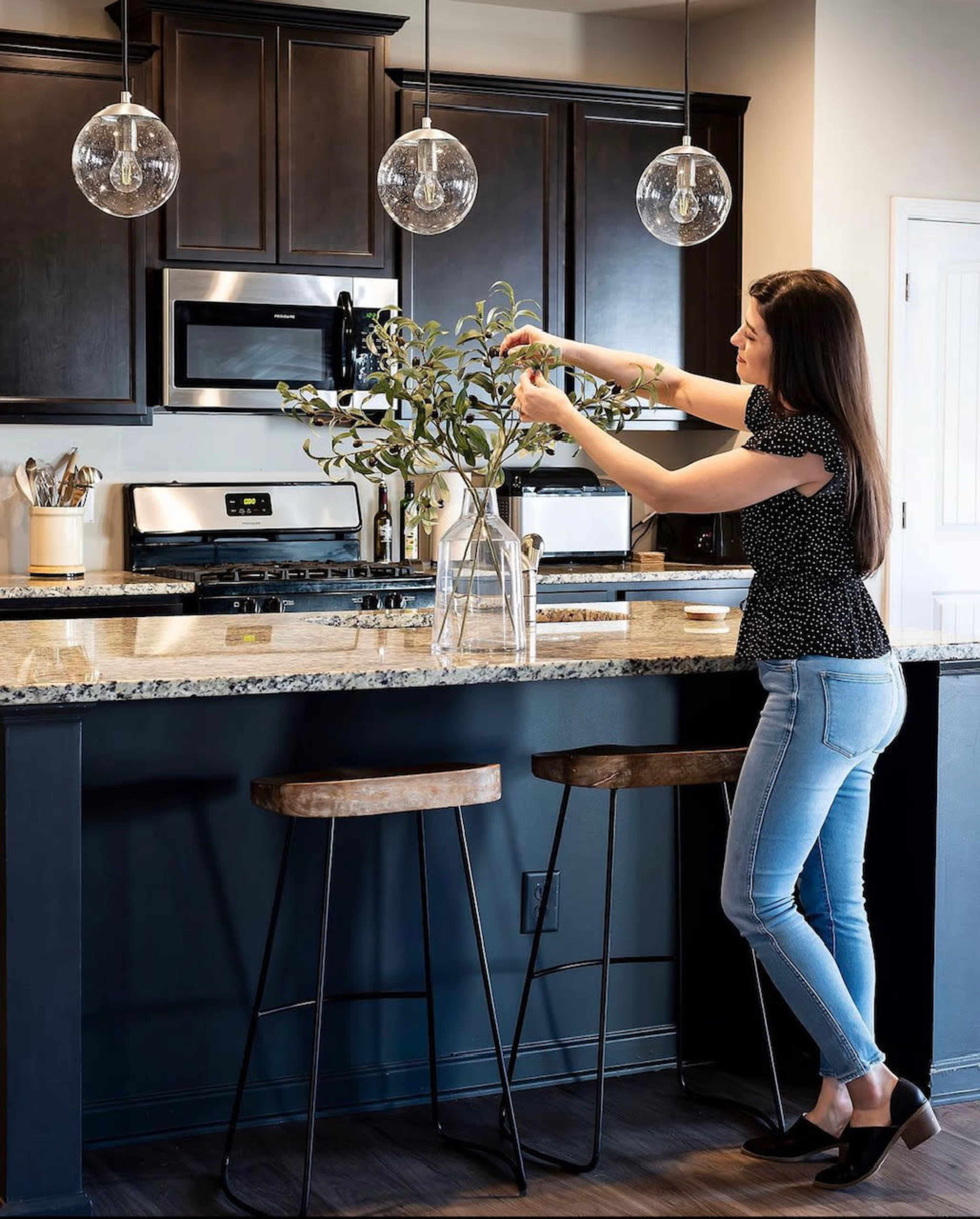 A woman arranges greenery in a glass vase on a kitchen island with dark wood cabinets and pendant lighting.