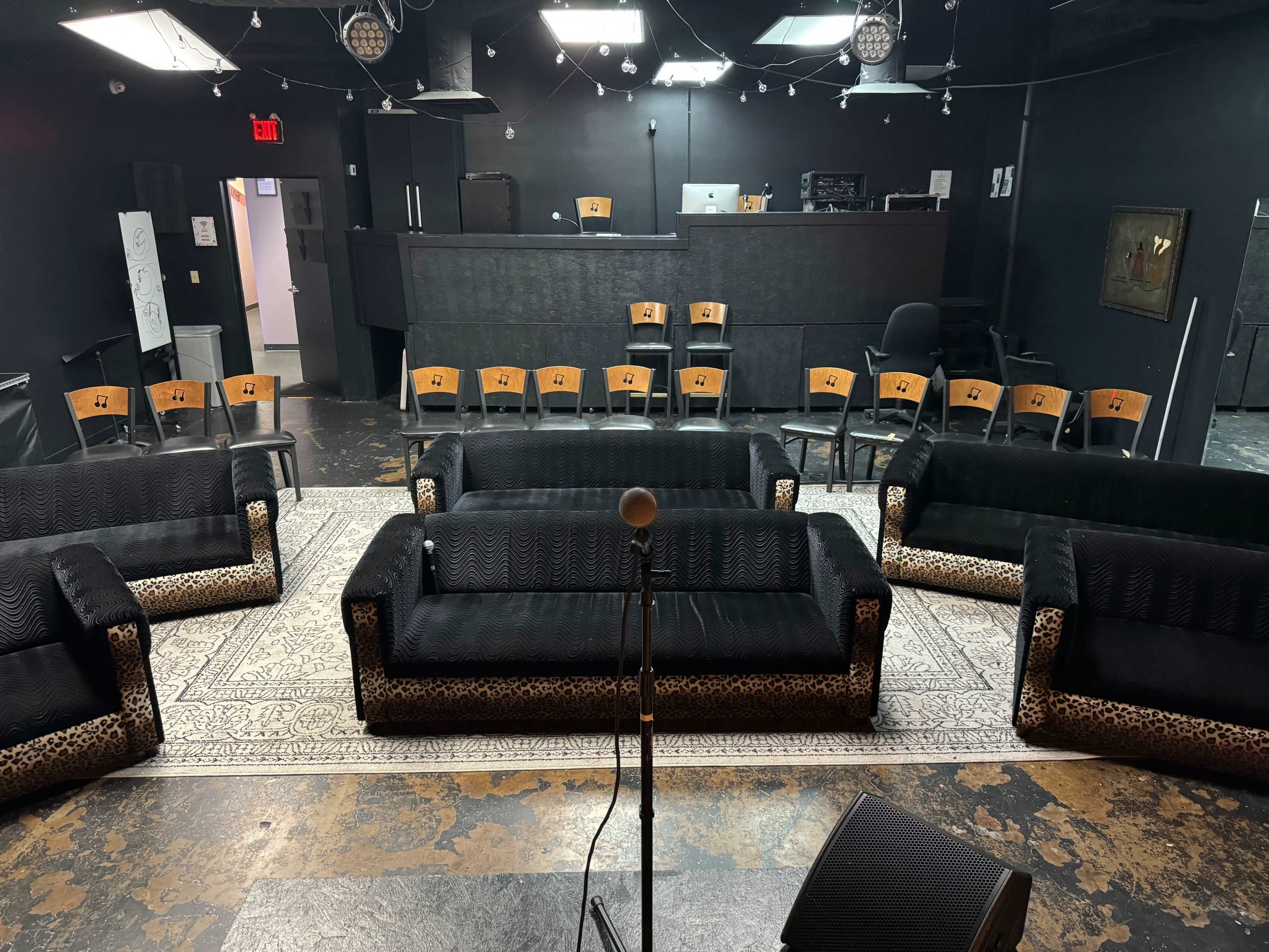 A dimly lit venue with several black couches arranged in a semicircle facing a microphone, and a row of chairs positioned in the background.