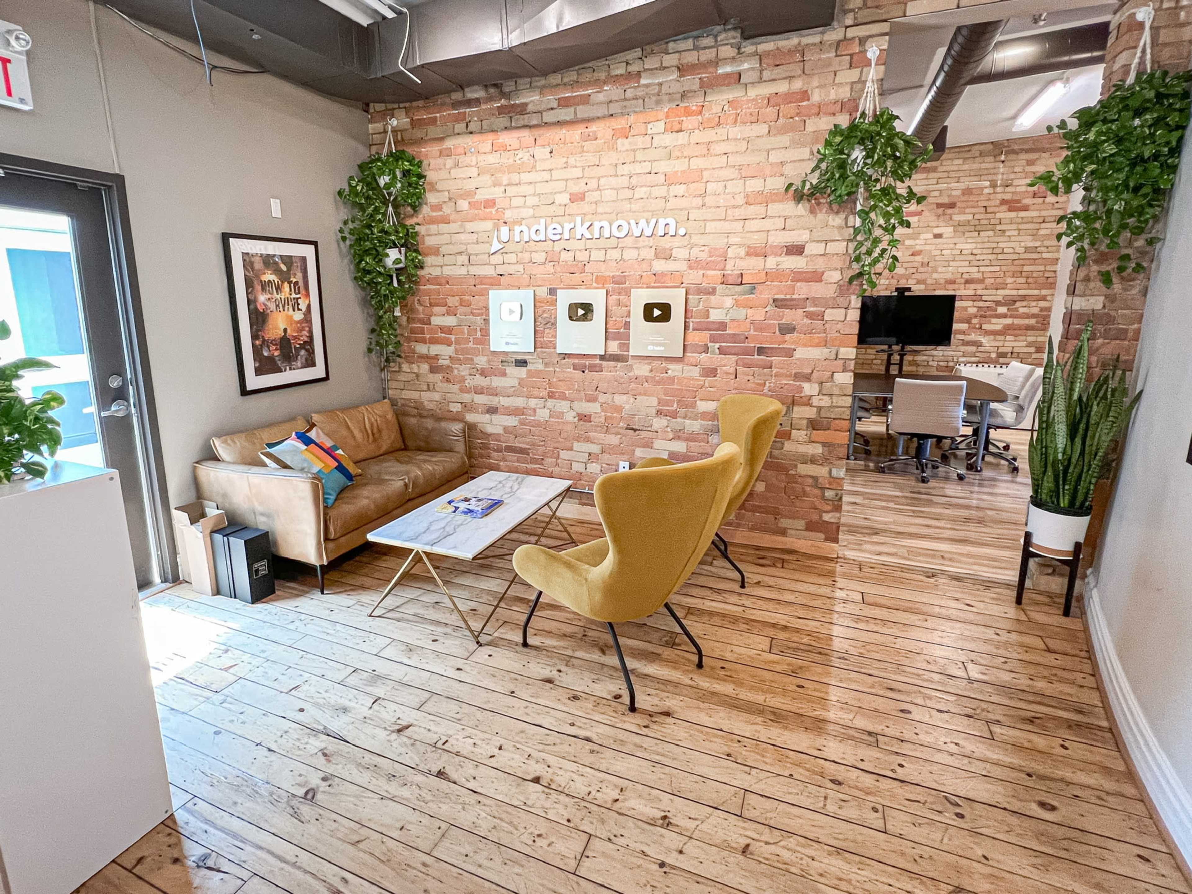 A modern office space with a leather couch, yellow chairs, a coffee table, and a wall decorated with hanging plants and plaques.