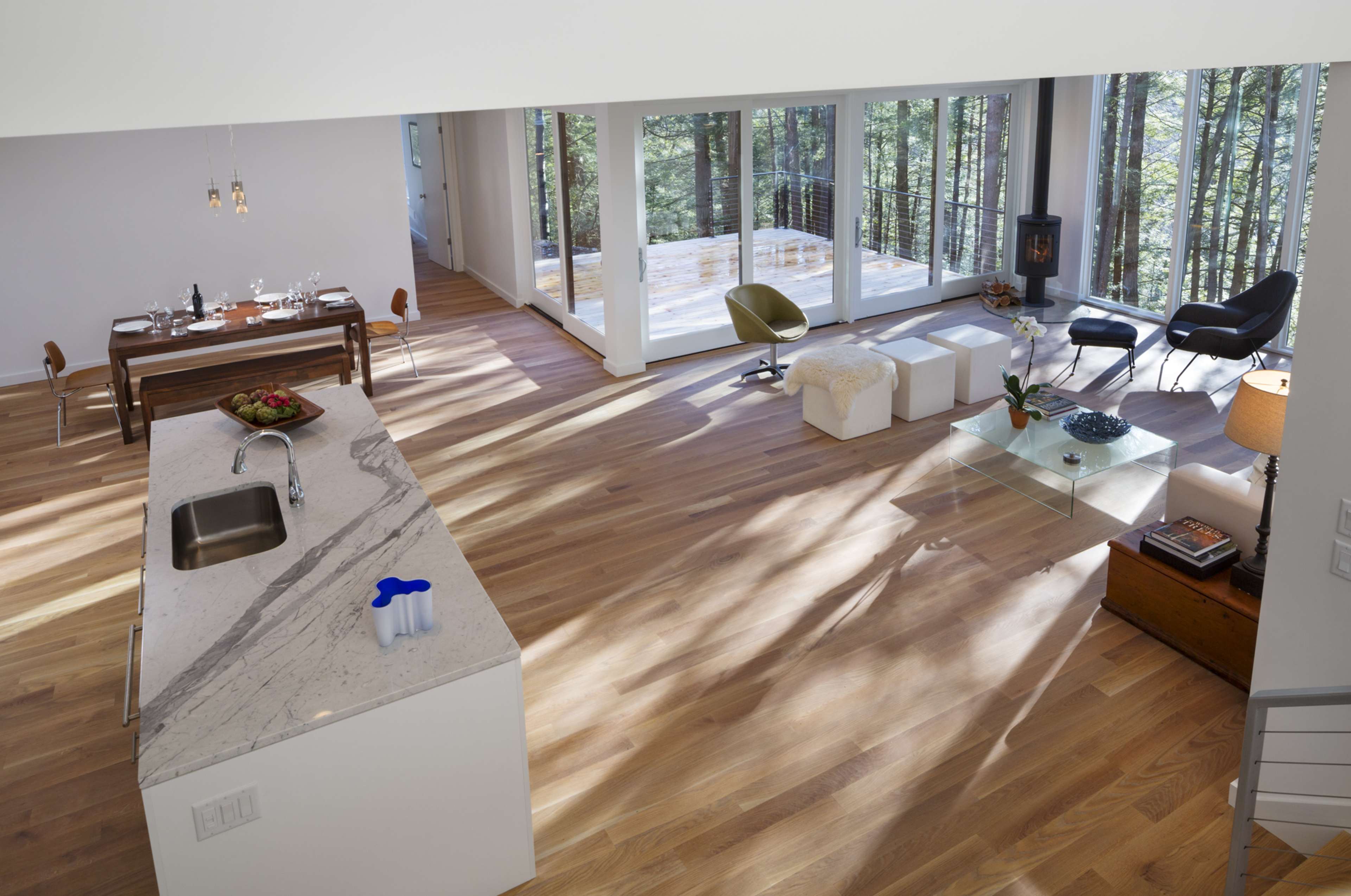 The image shows a spacious, modern interior with a kitchen island, dining area, and living space surrounded by large windows overlooking a forest.