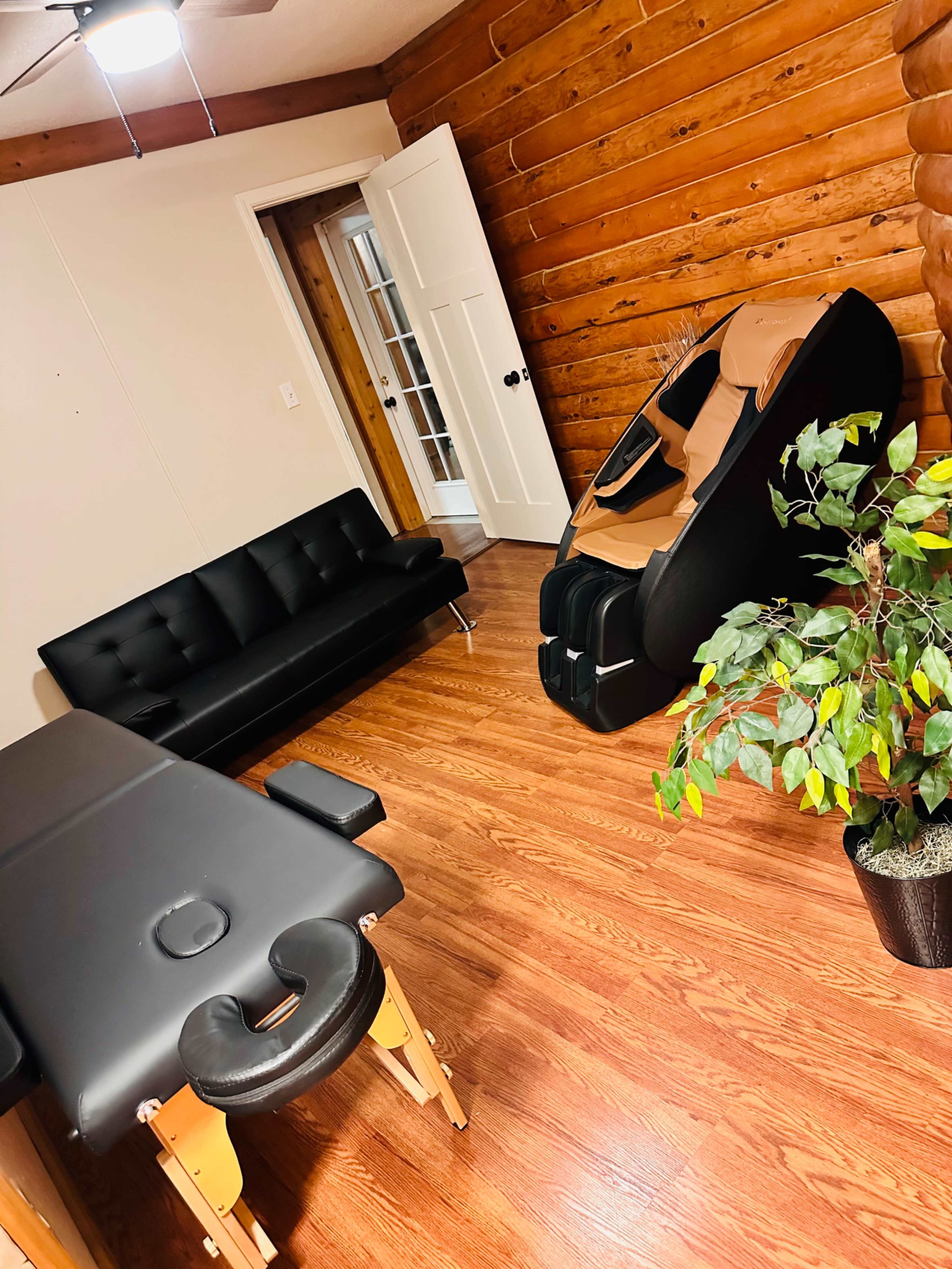 A room with wooden log walls, featuring a black sofa, a massage table, a massage chair, and a potted plant.