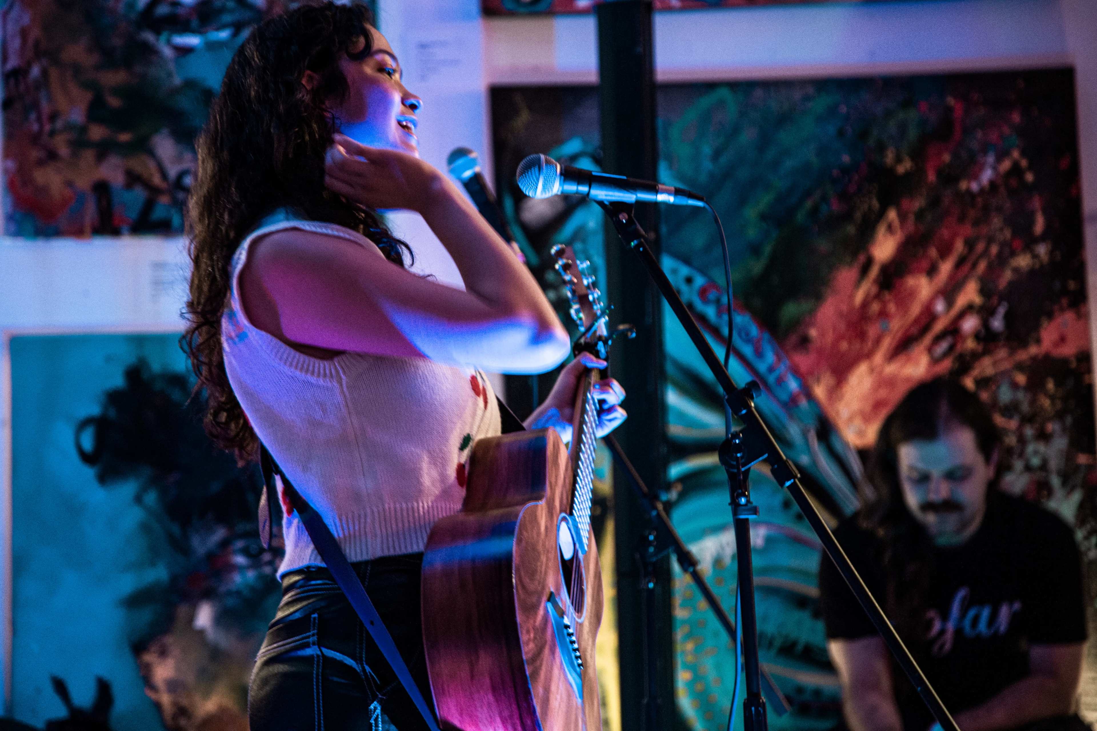 A woman sings into a microphone while holding an acoustic guitar, with a man playing an instrument in the background amidst colorful artwork.