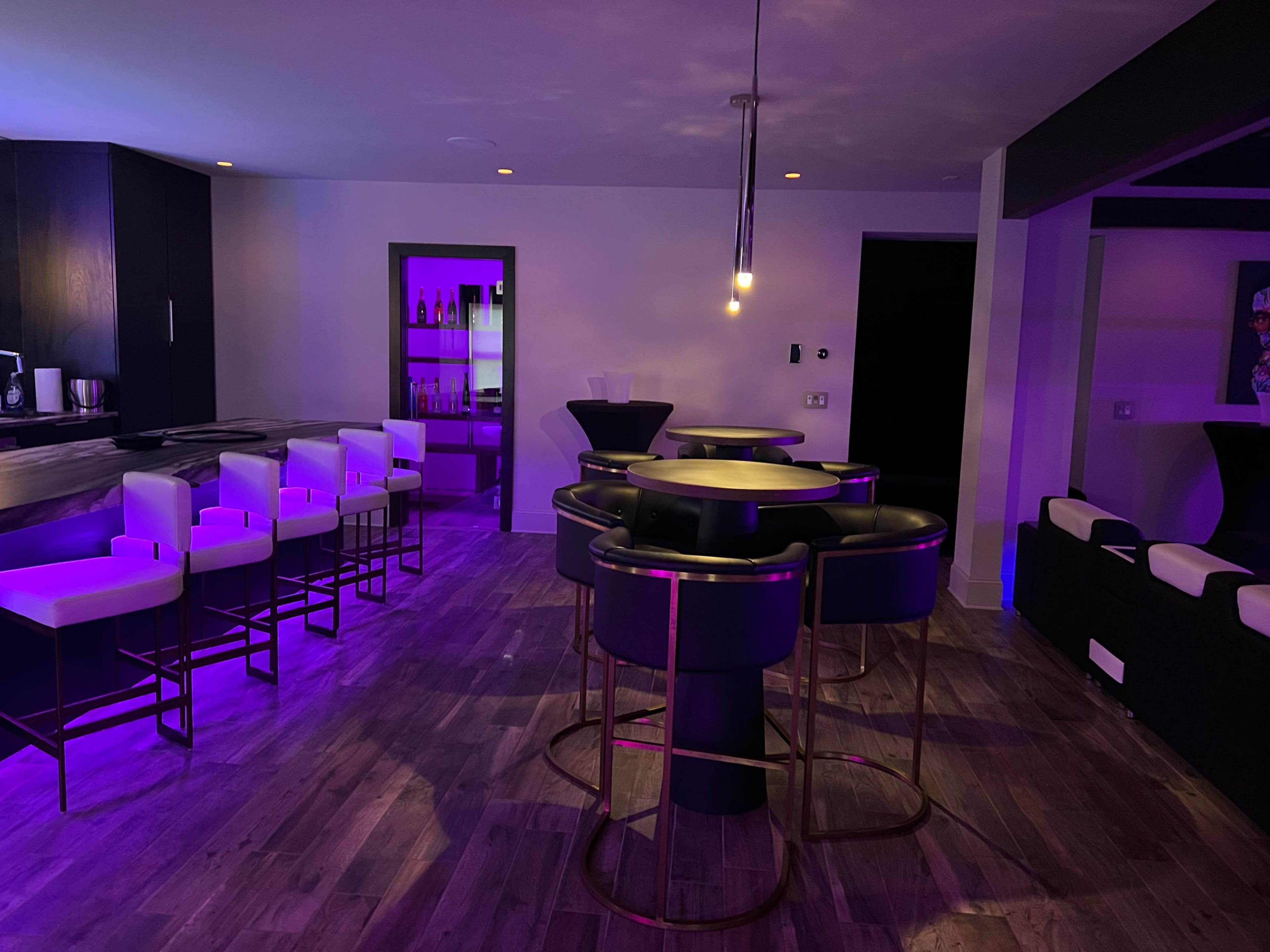 A modern lounge area features dark cabinetry, high bar stools with white seats, and purple ambient lighting.