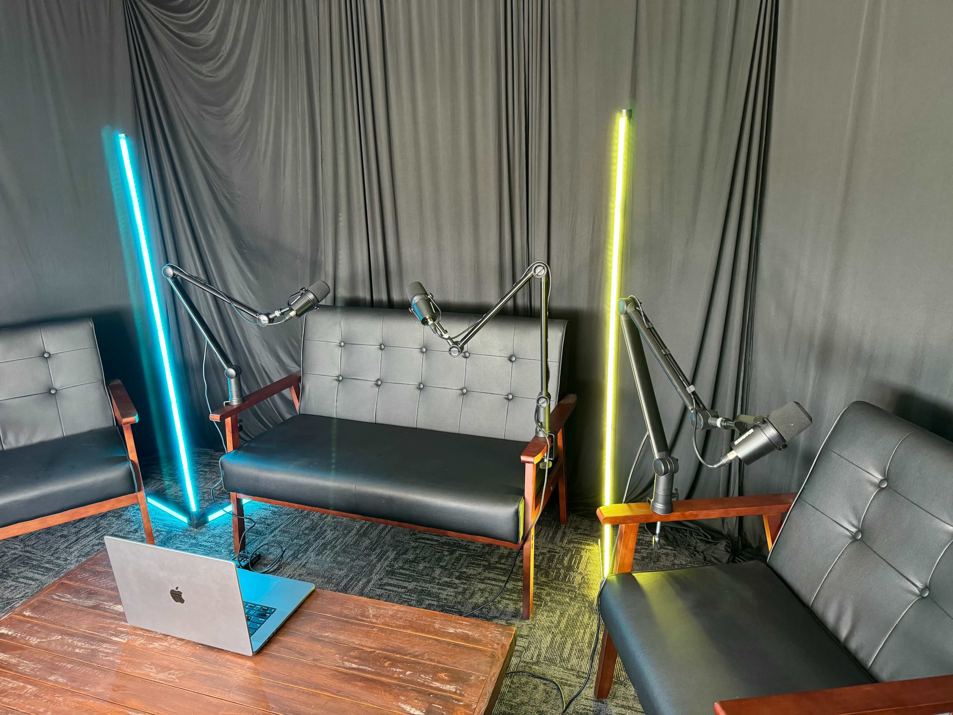 A recording setup with two black leather couches, two adjustable microphones on stands, and a laptop on a wooden table, all set against a backdrop of dark fabric.