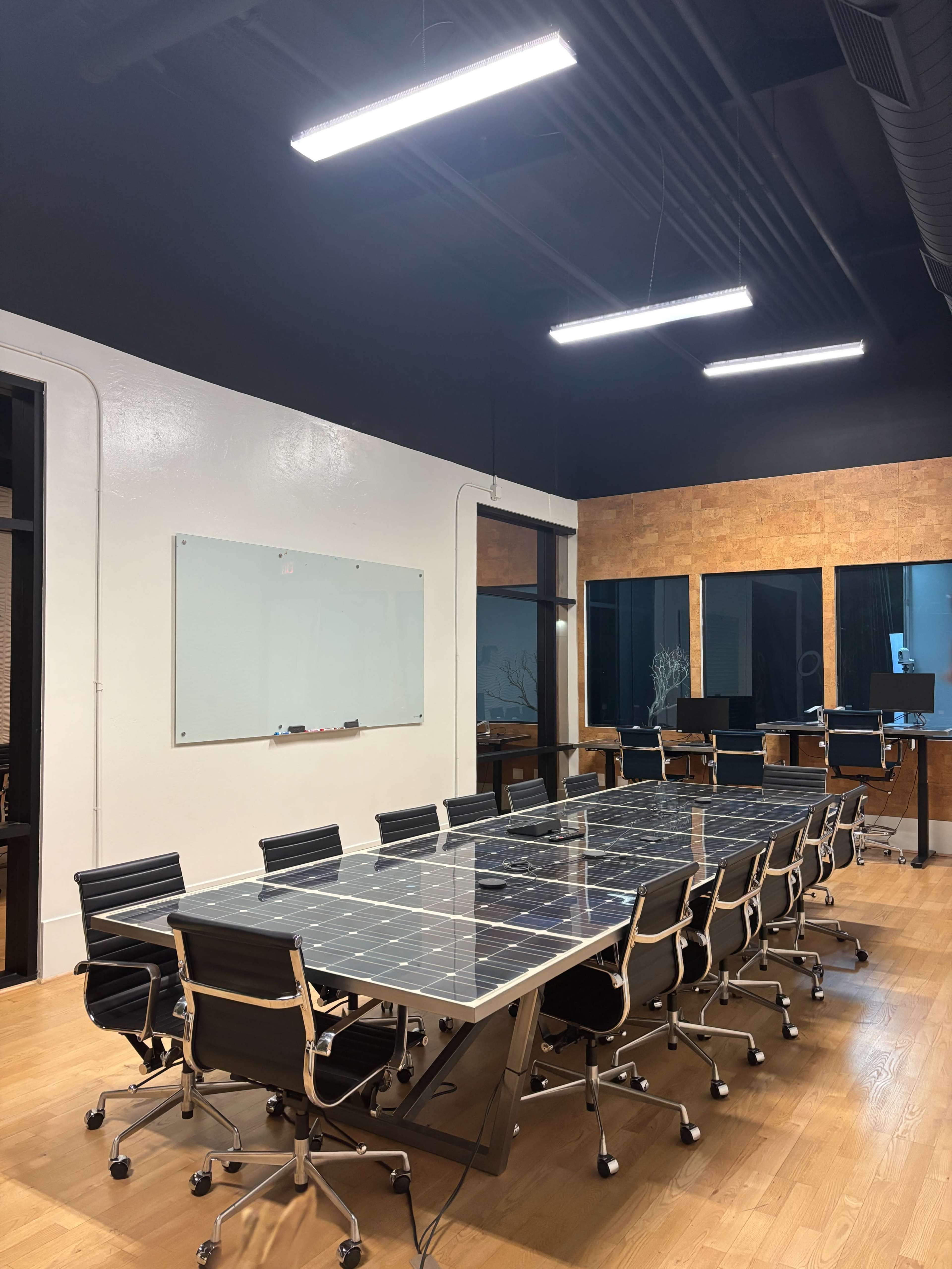 A modern conference room features a large table with a glass top surrounded by black chairs, illuminated by overhead lights.