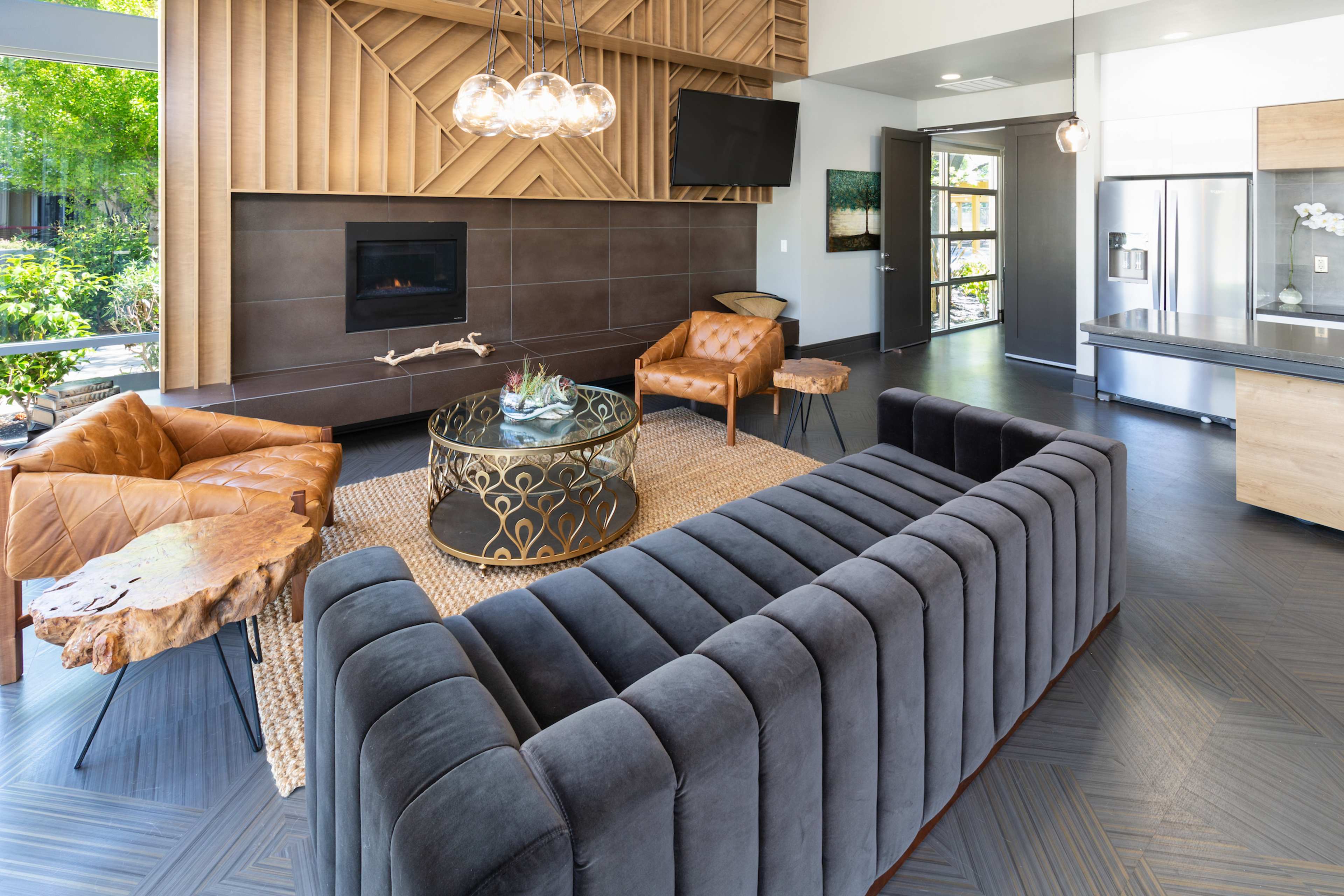 A modern living room featuring a grey sectional sofa, a round coffee table with a decorative centerpiece, two tan armchairs, and a wall-mounted television above a minimalistic fireplace.