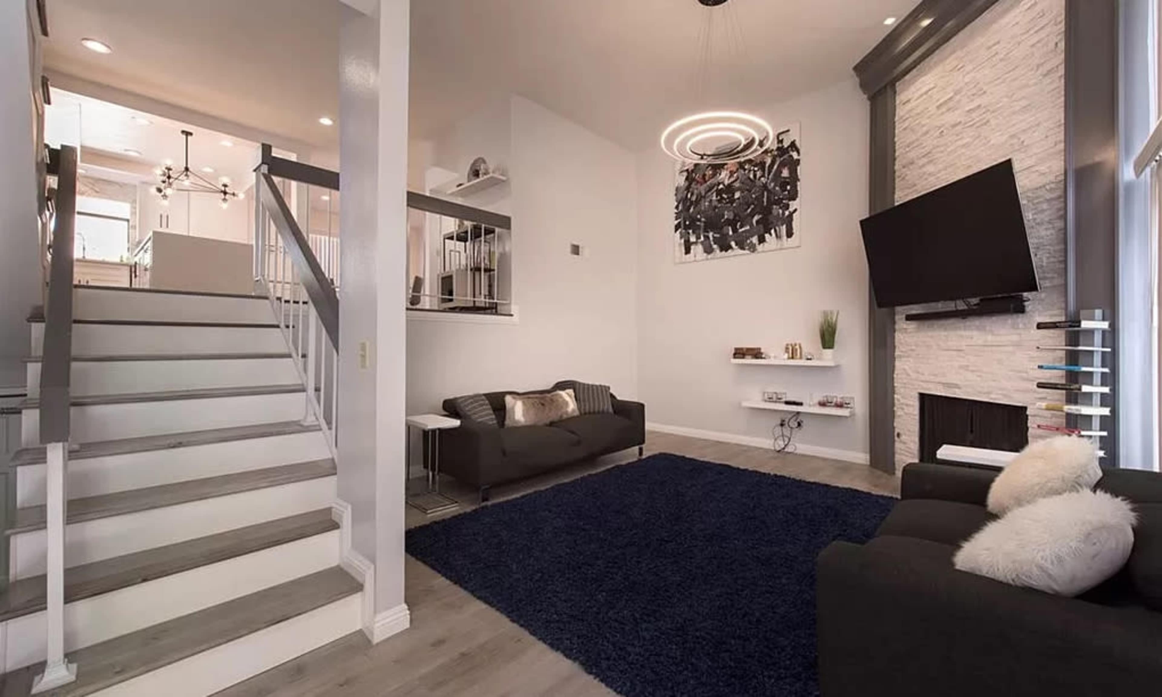 A modern living room features a staircase, a large dark rug, a sectional sofa, a wall-mounted television, and a decorative wall with a circular light fixture.