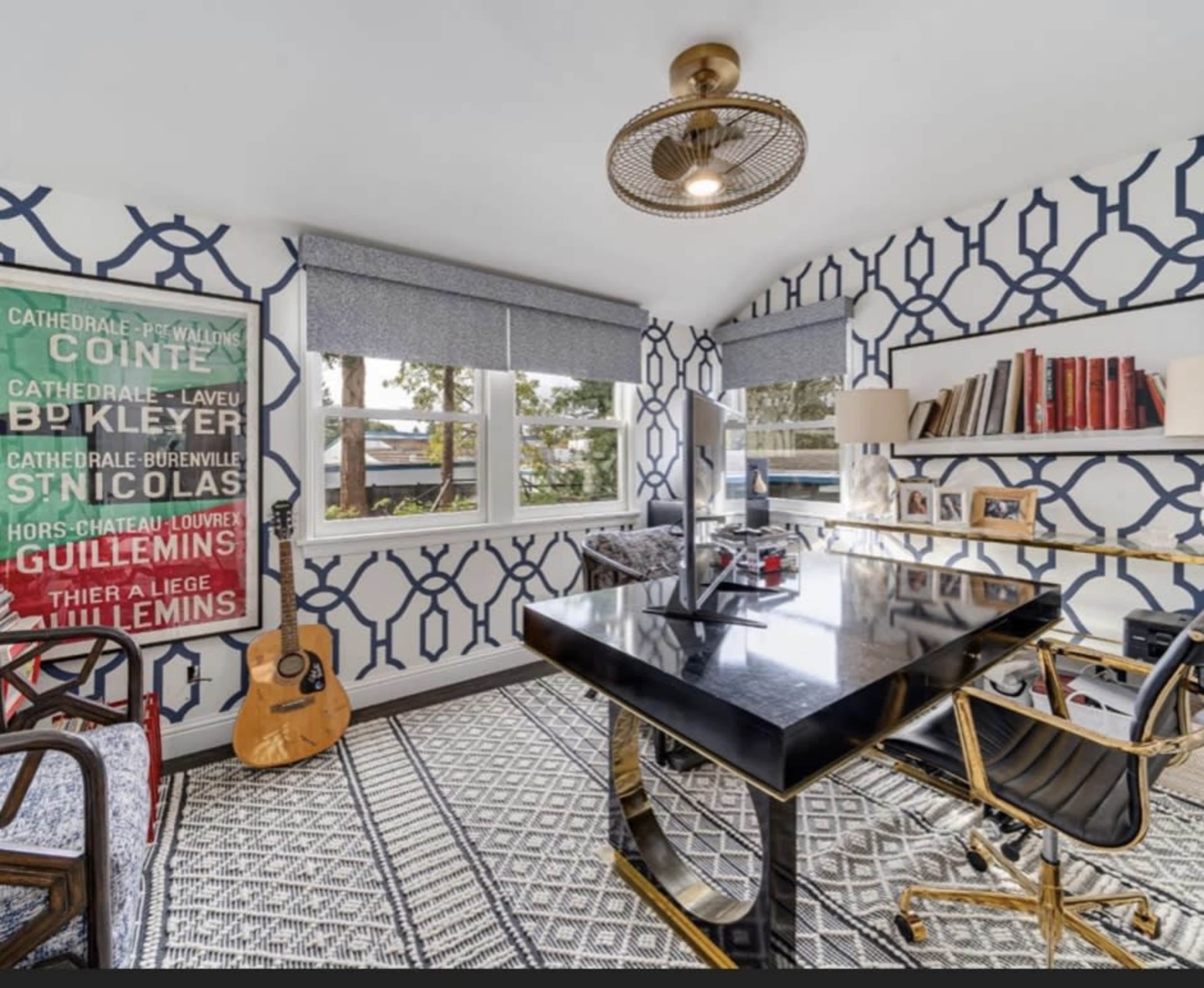 A modern home office featuring a black desk, geometric-patterned wallpaper, a guitar leaning against the wall, and large windows allowing natural light.