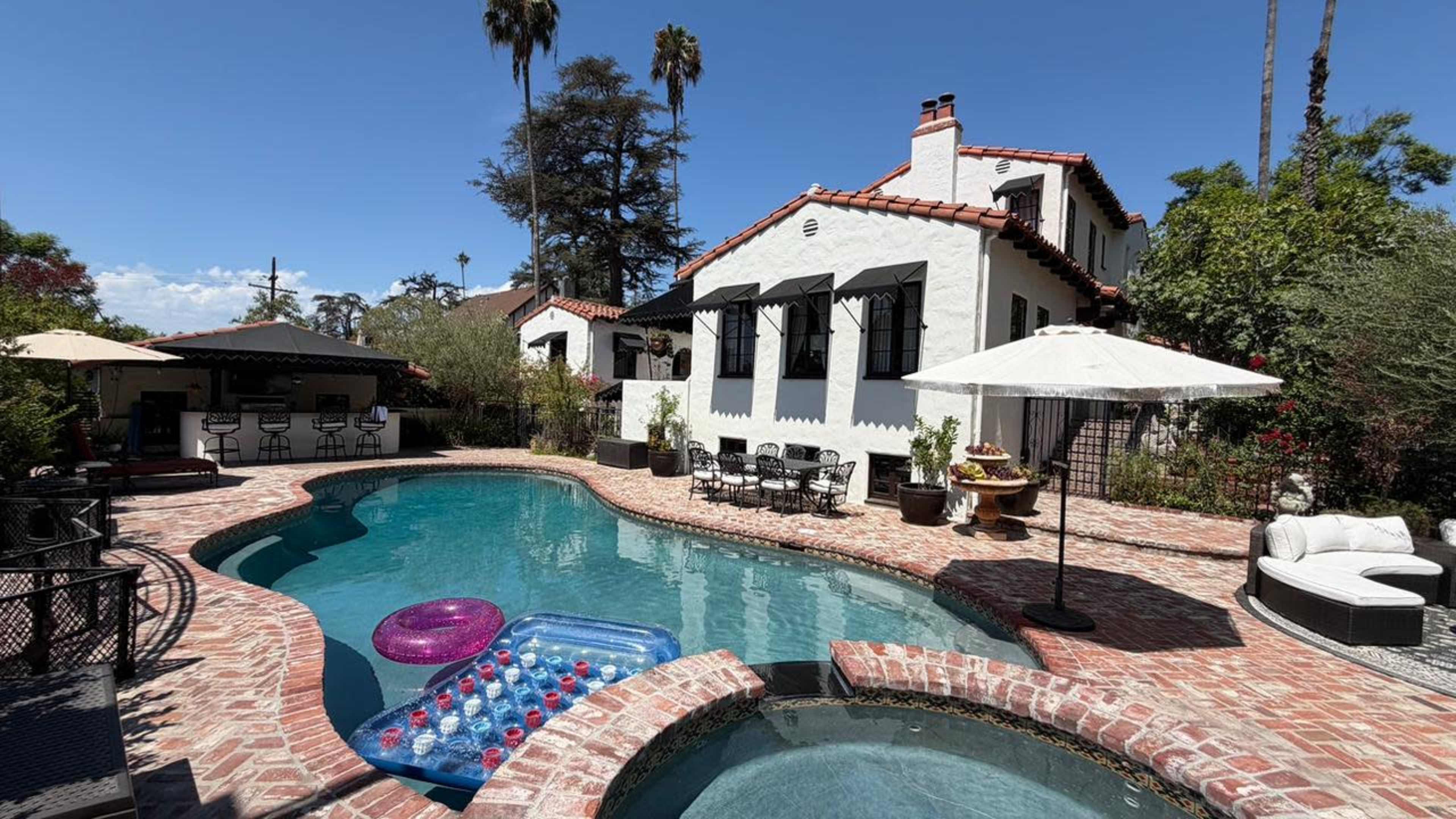 The image shows a swimming pool surrounded by brick paving, with lounge chairs, inflatable pool toys, and a house featuring a tiled roof and palm trees in the background.