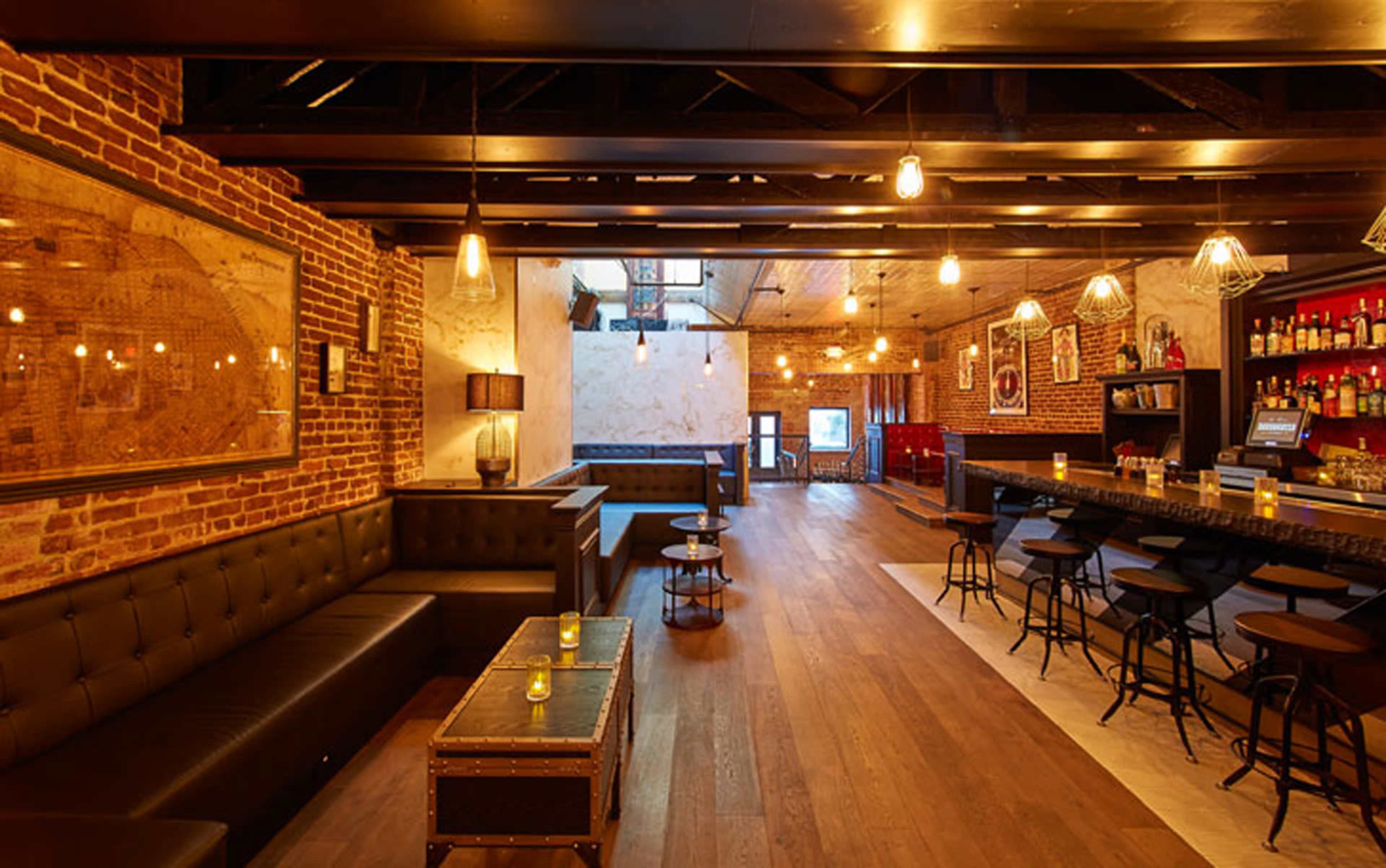 The image depicts a bar interior featuring exposed brick walls, wooden flooring, and a long bar with high stools, illuminated by pendant lighting.