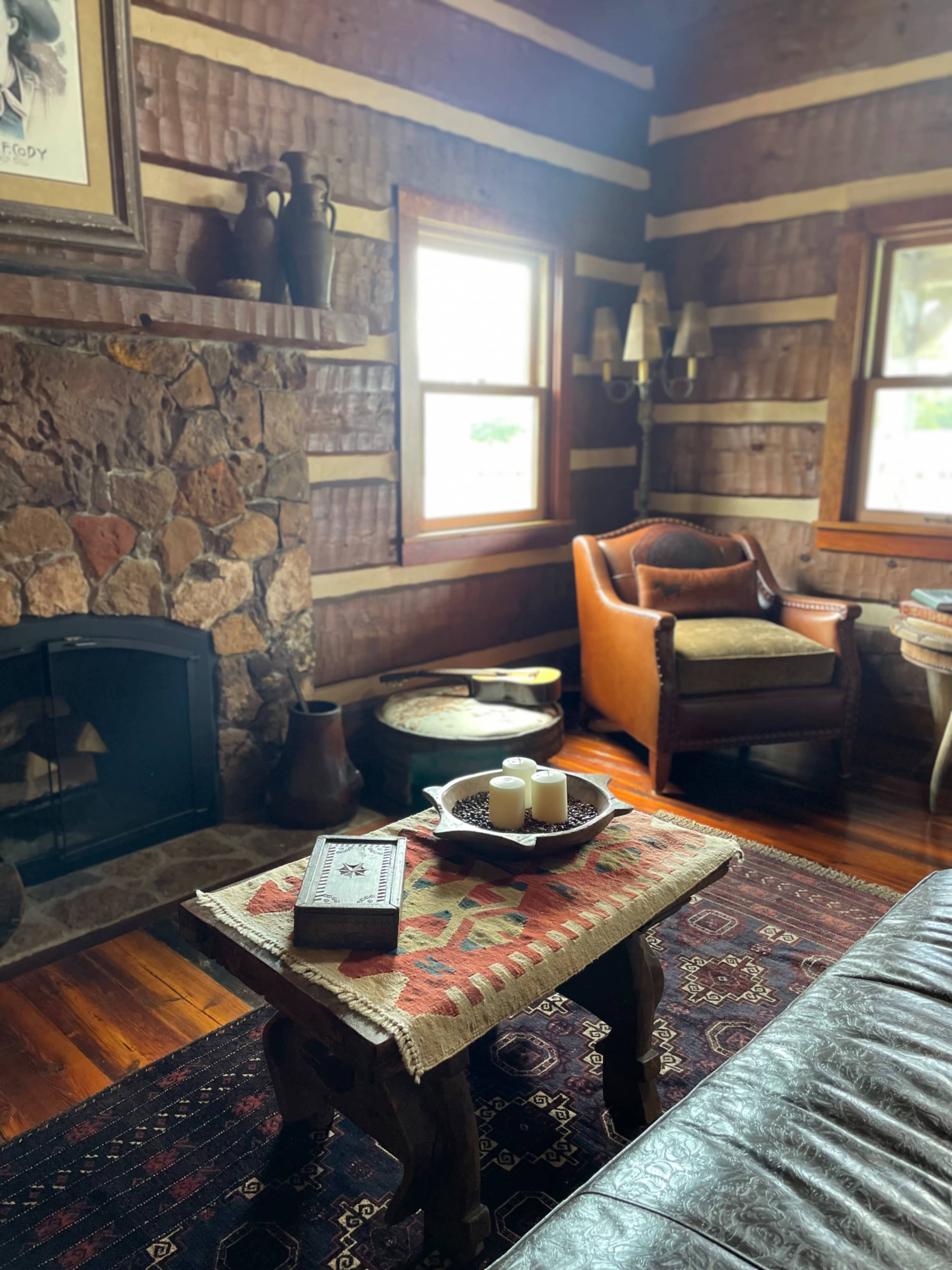 The image shows a rustic log cabin living room featuring a stone fireplace, wooden furniture, and a patterned rug.
