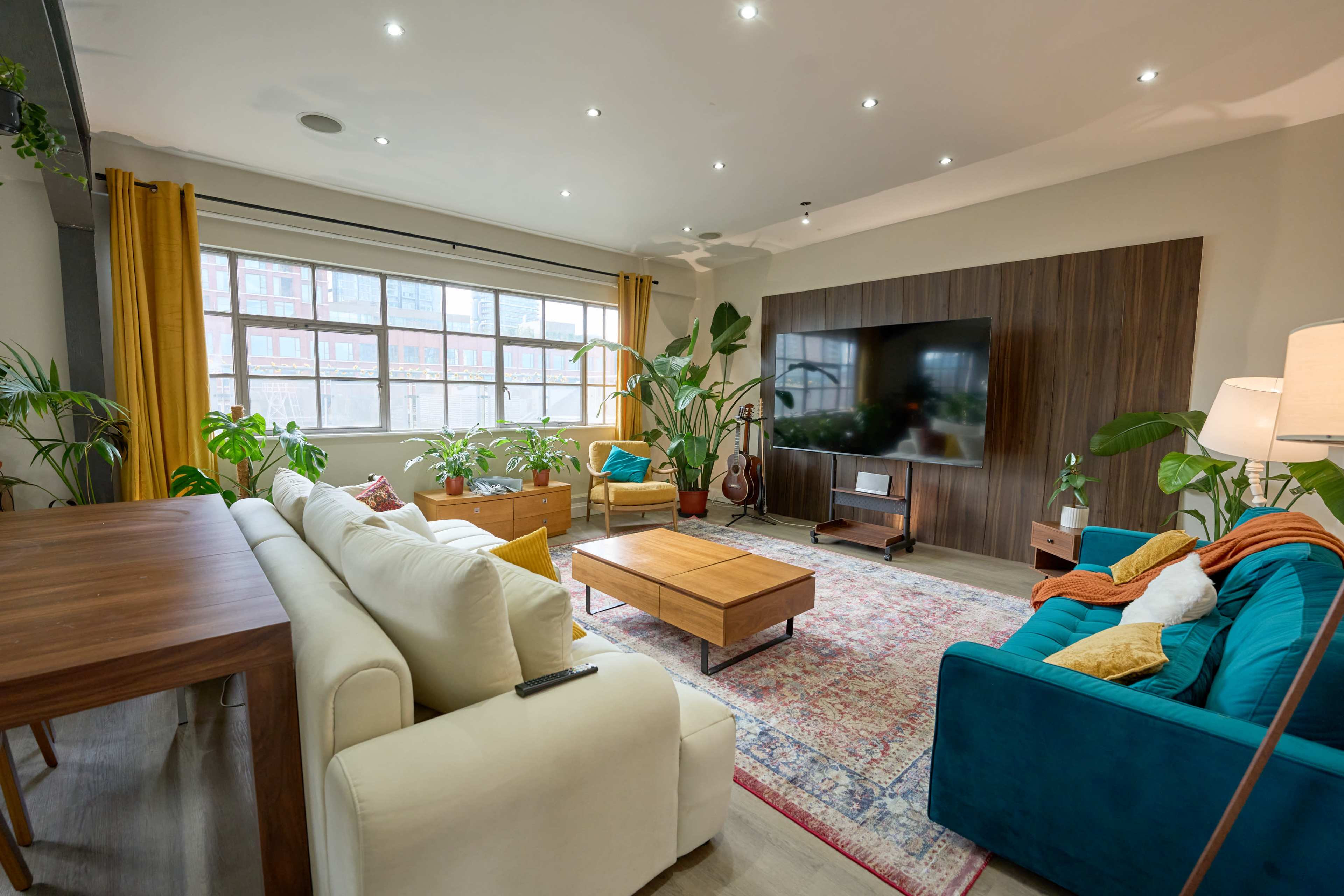 The image features a modern living room with a sectional sofa, wooden coffee table, large TV mounted on a wood-paneled wall, and various indoor plants.
