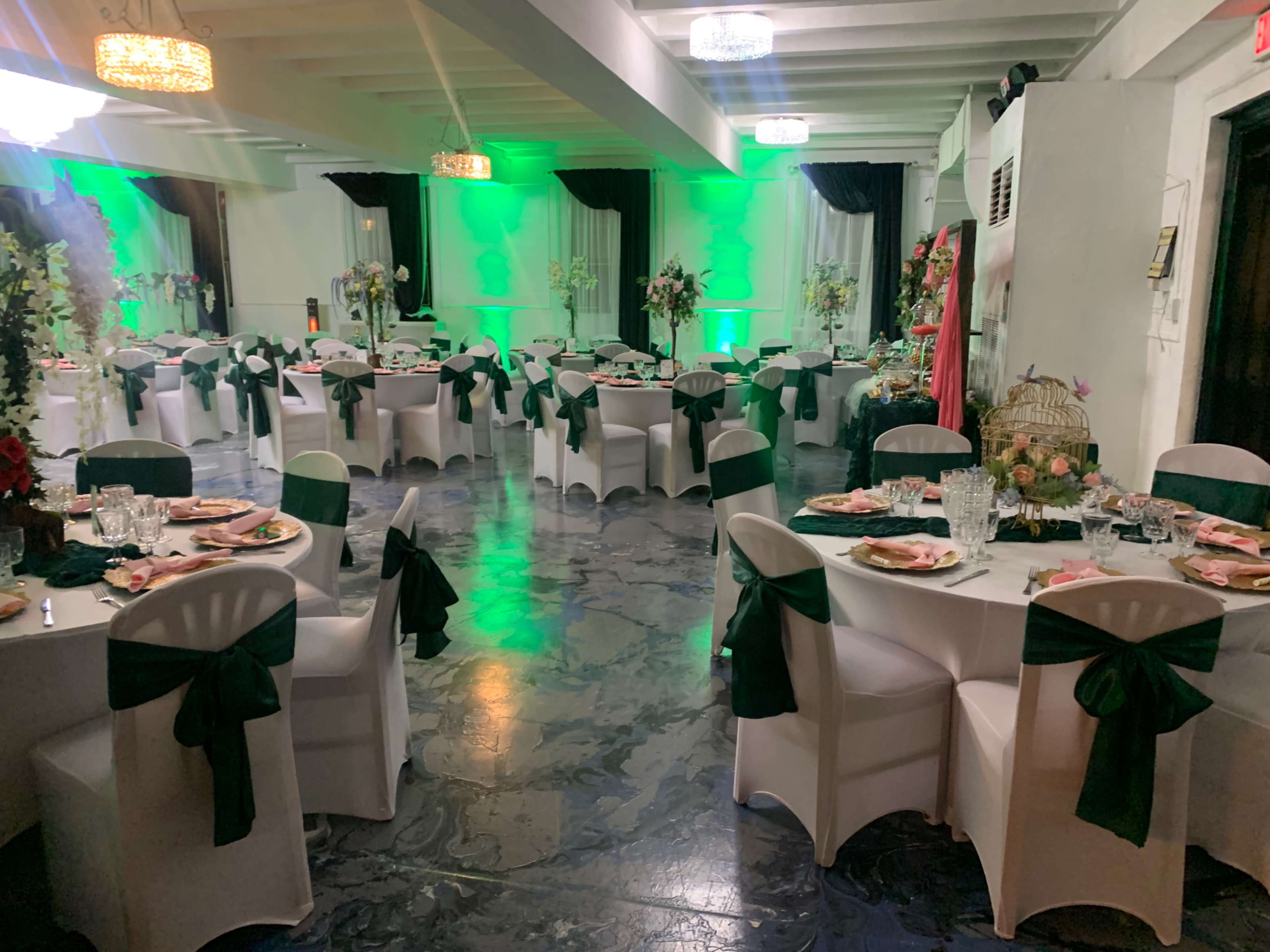 A banquet hall features round tables set with dark green bows on white chairs, surrounded by decorative floral arrangements and illuminated by green ambient lighting.