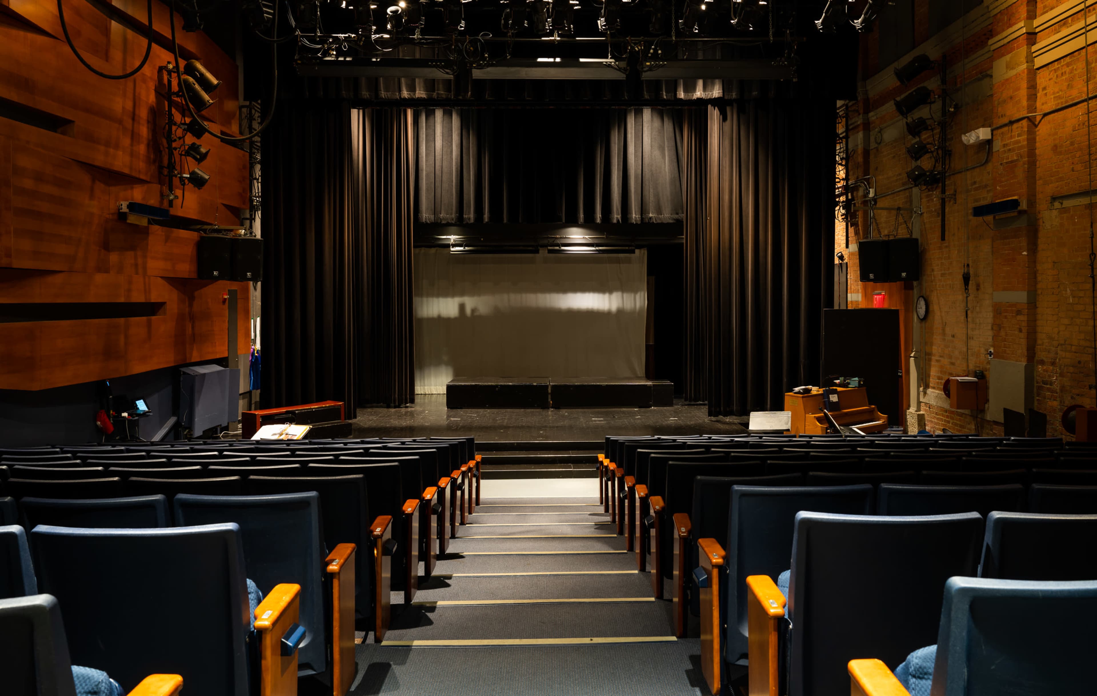 An empty theater with rows of blue seats facing a dark stage draped in black curtains.