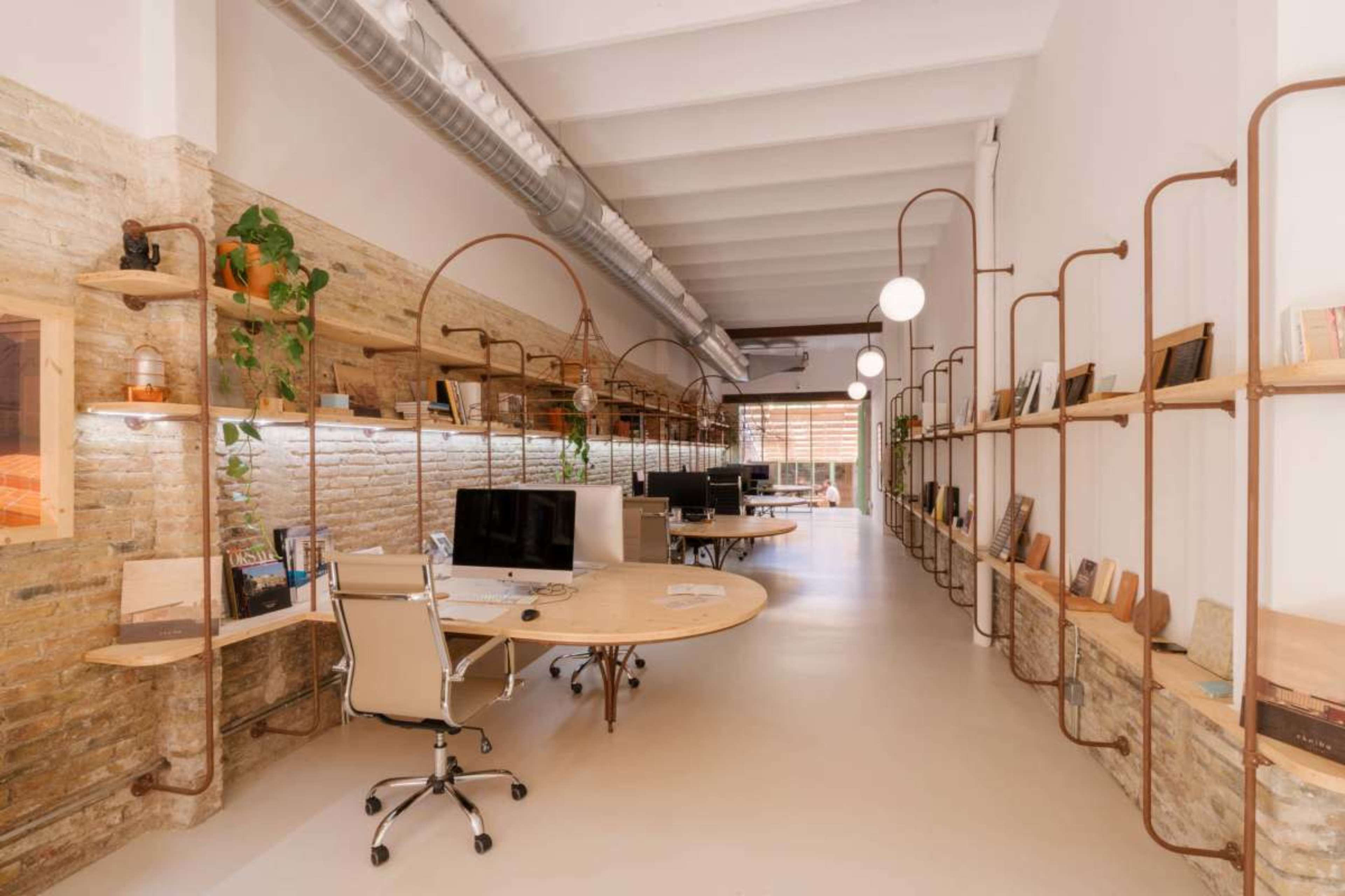 The image shows a modern office space with a long, open layout featuring desks, computers, and shelves along the walls.