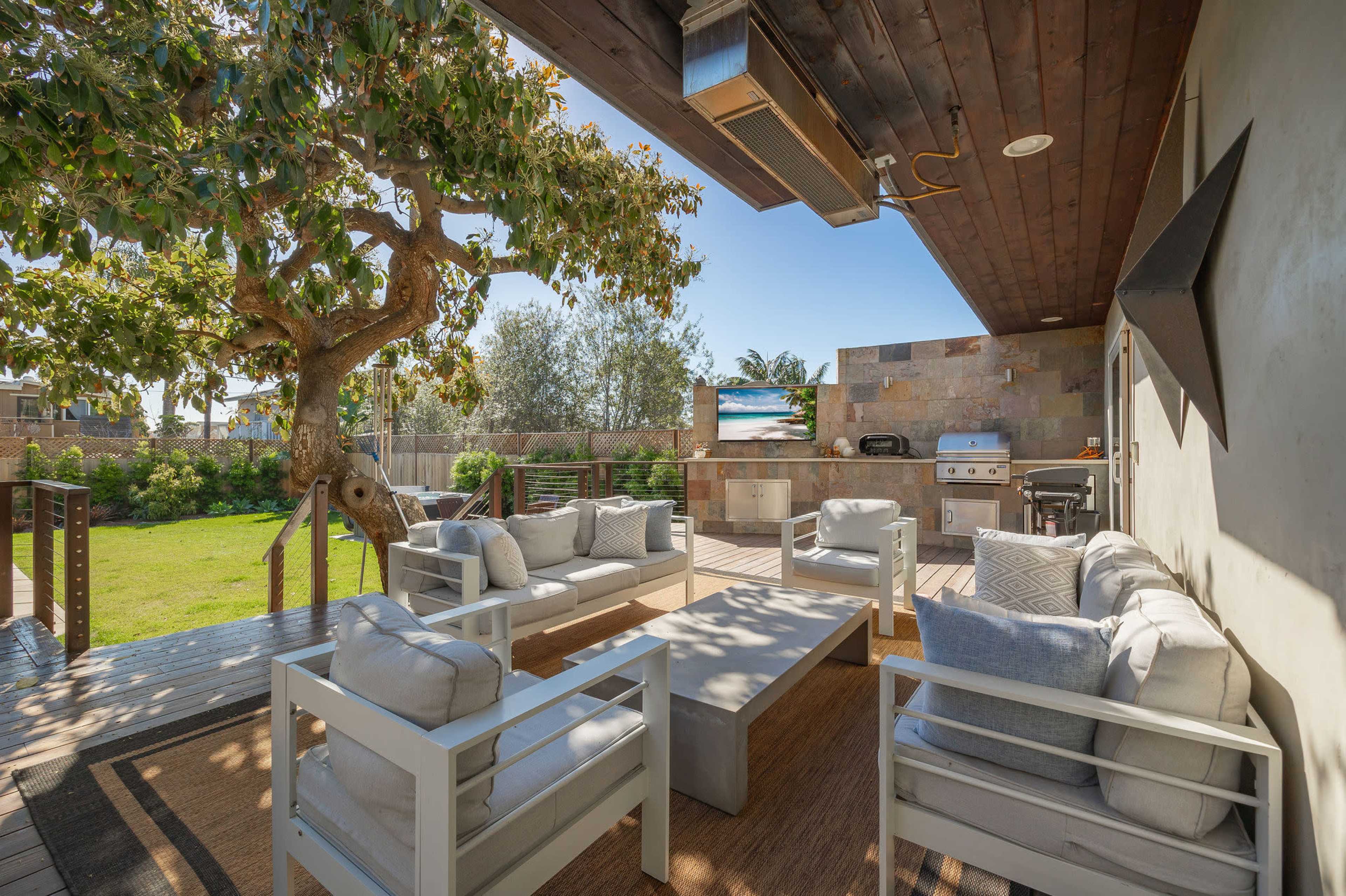 The image shows a patio area with white outdoor furniture, a barbecue grill, and a large television mounted on a stone wall, surrounded by greenery.