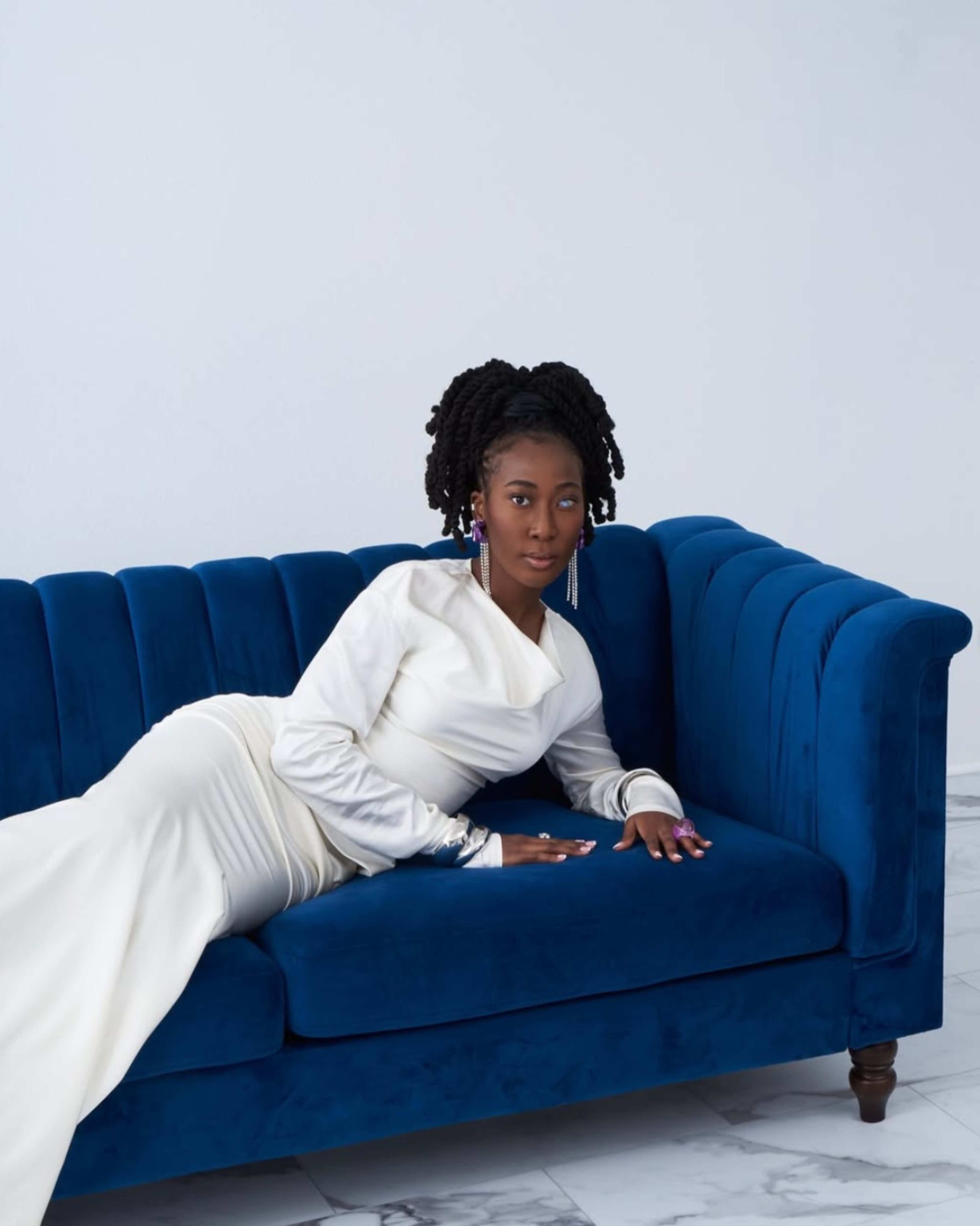 A woman in a white dress reclines on a blue velvet couch against a plain backdrop.