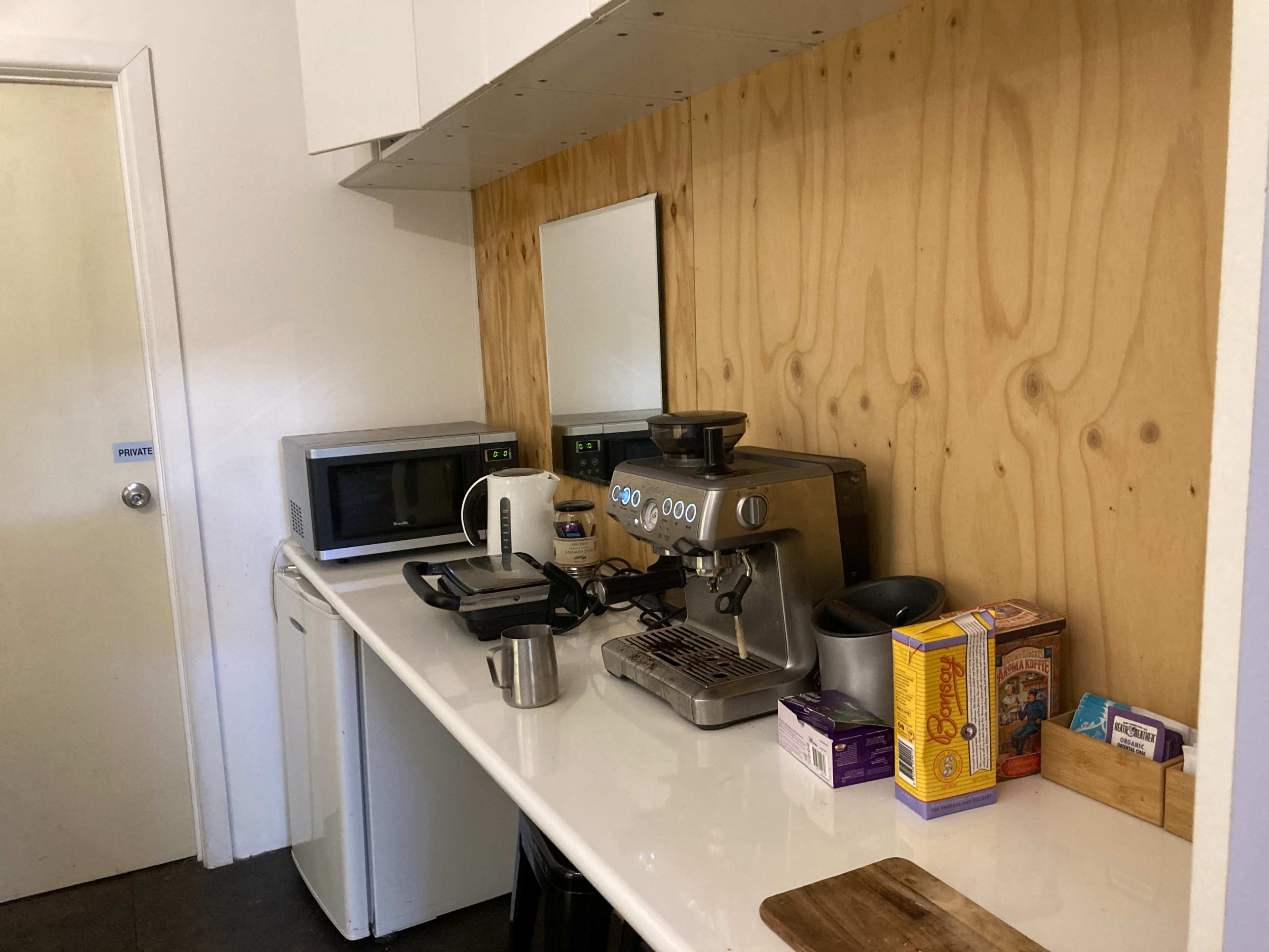 A compact kitchen features a coffee machine, microwave, and various food items neatly arranged on a countertop with a wooden back wall.