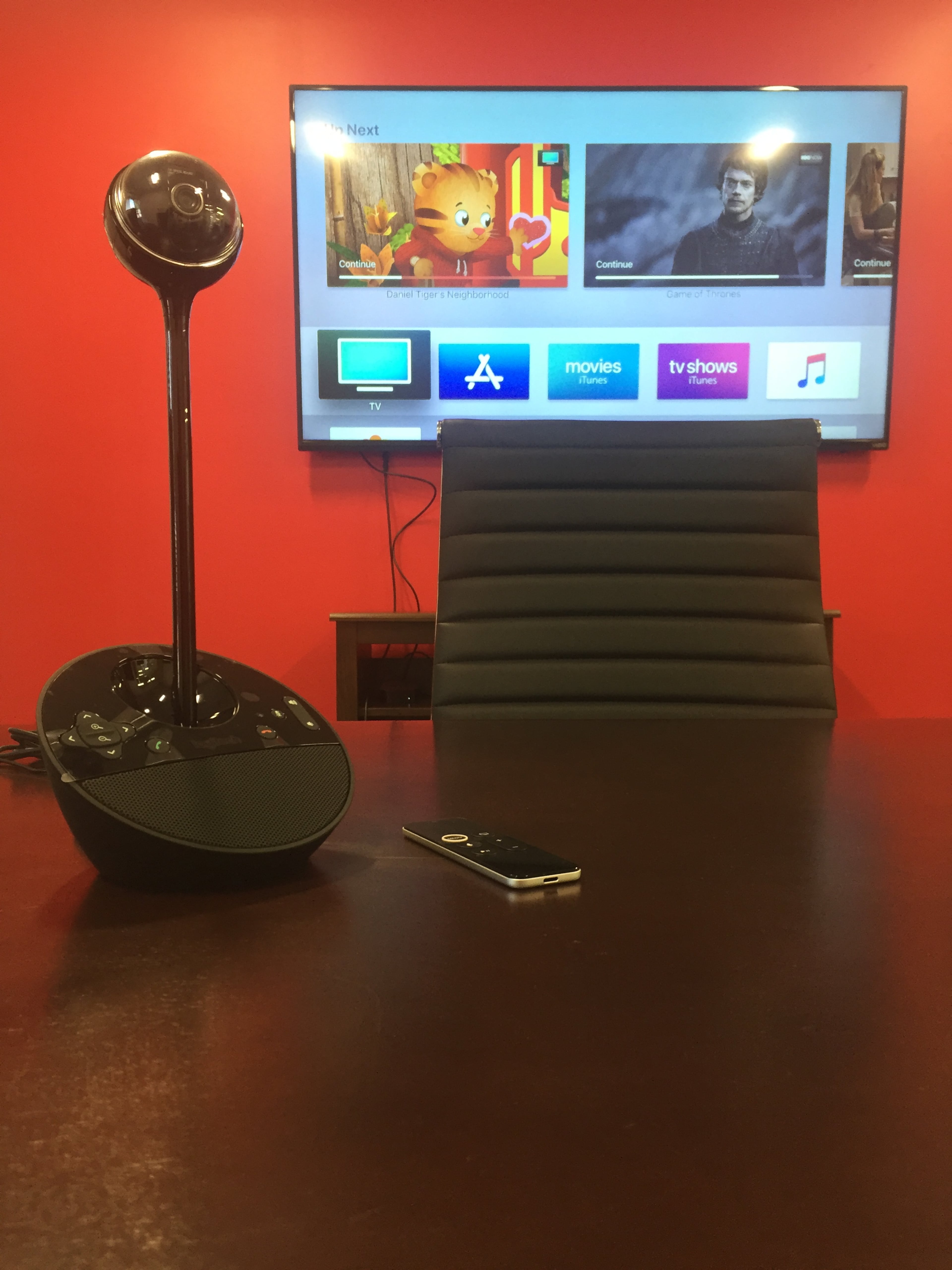 A conference room features a sleek black chair and a control device on the table in front of a television mounted on a red wall.