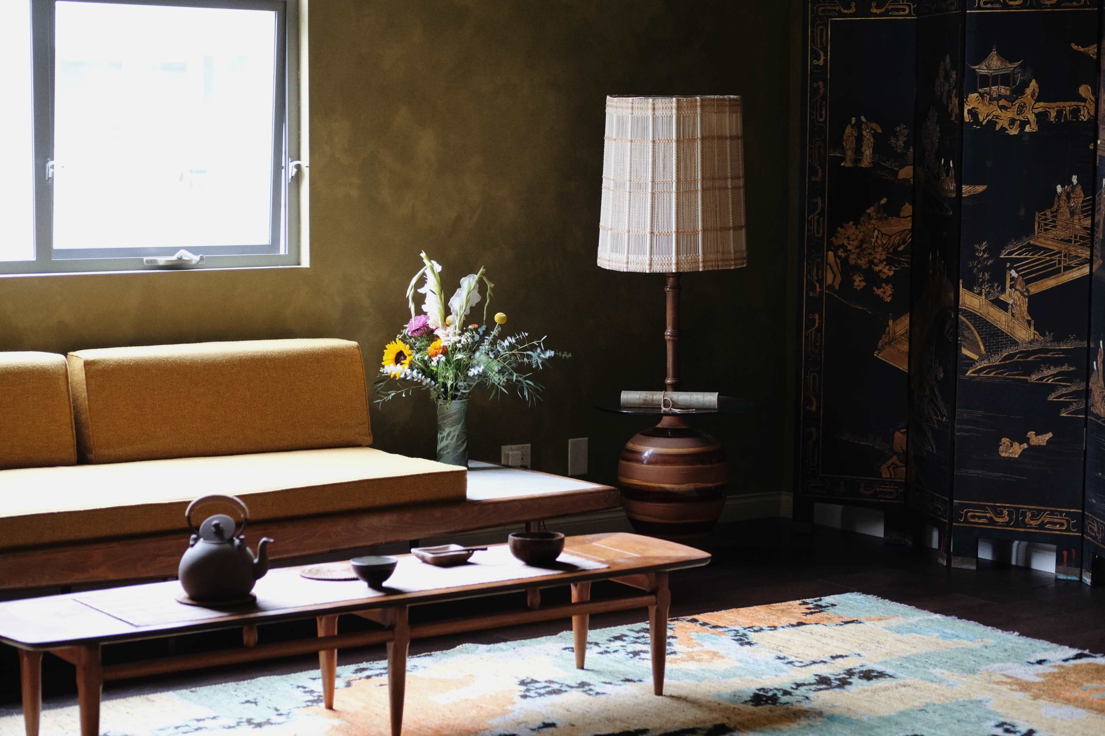 The image shows a cozy living room with a yellow sofa, a wooden coffee table with tea items, a patterned rug, a floor lamp, and a decorative screen.