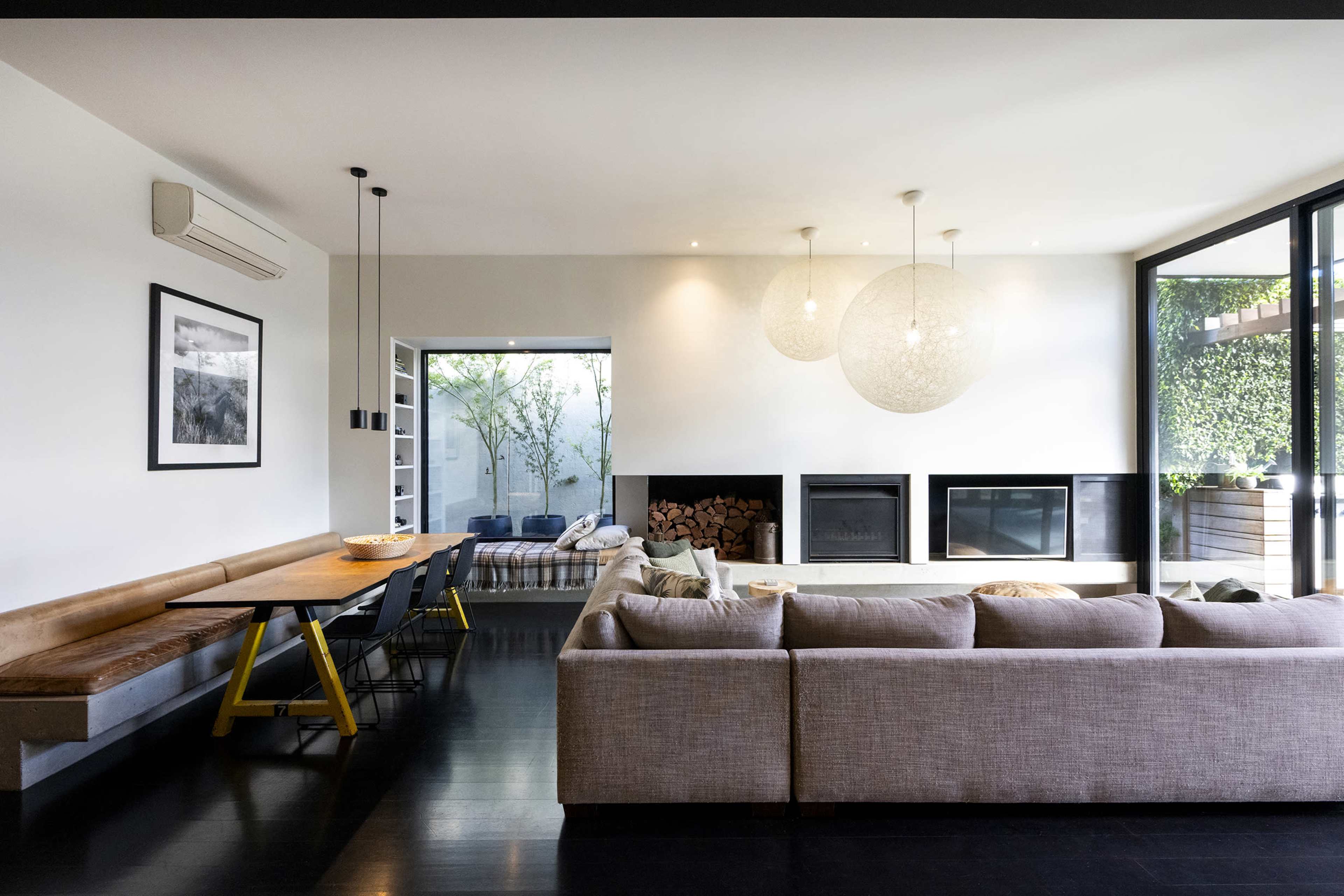 The image shows a modern living room featuring a large beige sofa, a wooden dining table with yellow legs, two fireplaces, and large windows that open to a green outdoor space.