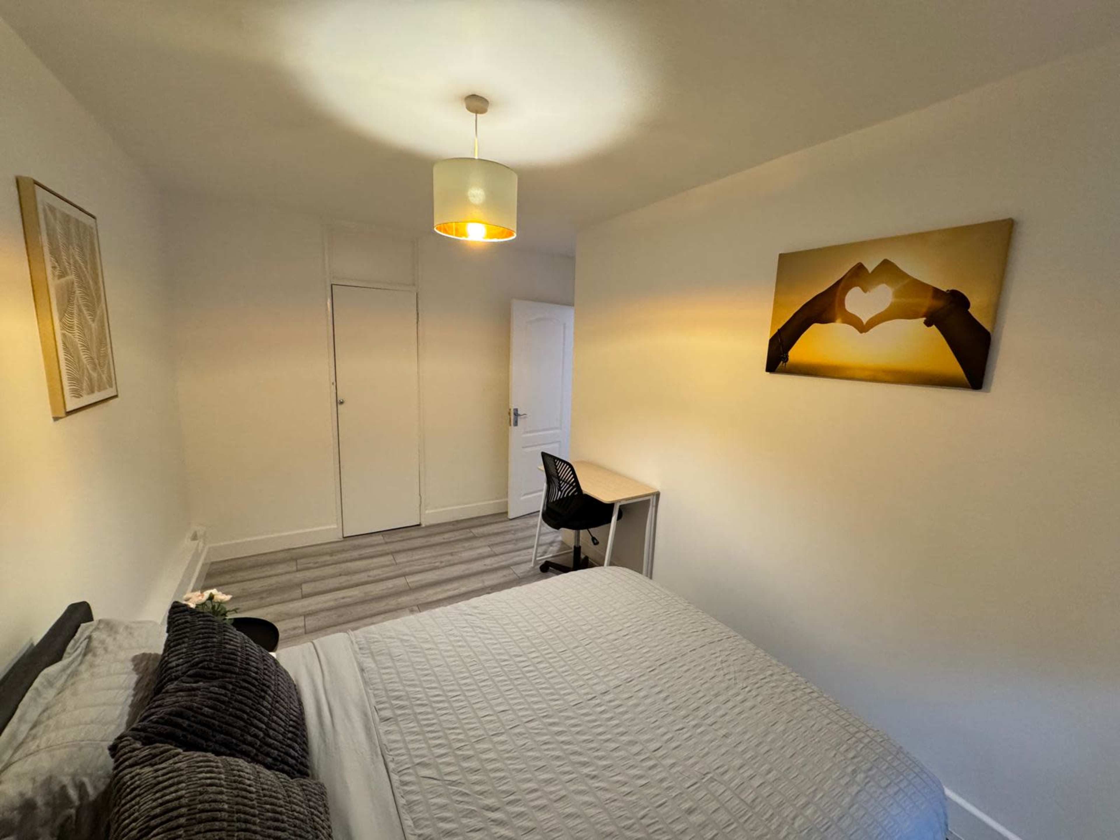 A well-lit bedroom features a bed with gray bedding, a wall-mounted artwork of hands forming a heart, a desk with a chair, and a light fixtures hanging from the ceiling.